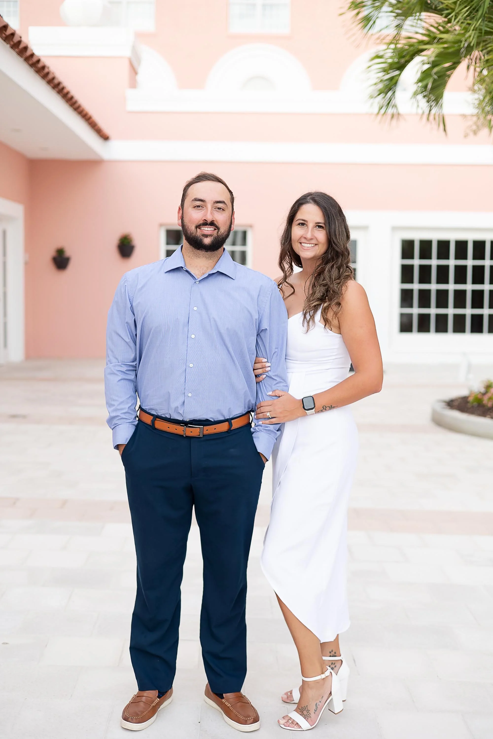 A man and woman standing together outside in front of a pink building with white accents. The man is wearing a light blue shirt, dark blue pants, and brown shoes. The woman is wearing a white dress and white high heels, with tattoos on her ankle and foot. They are smiling and posing for the photo.