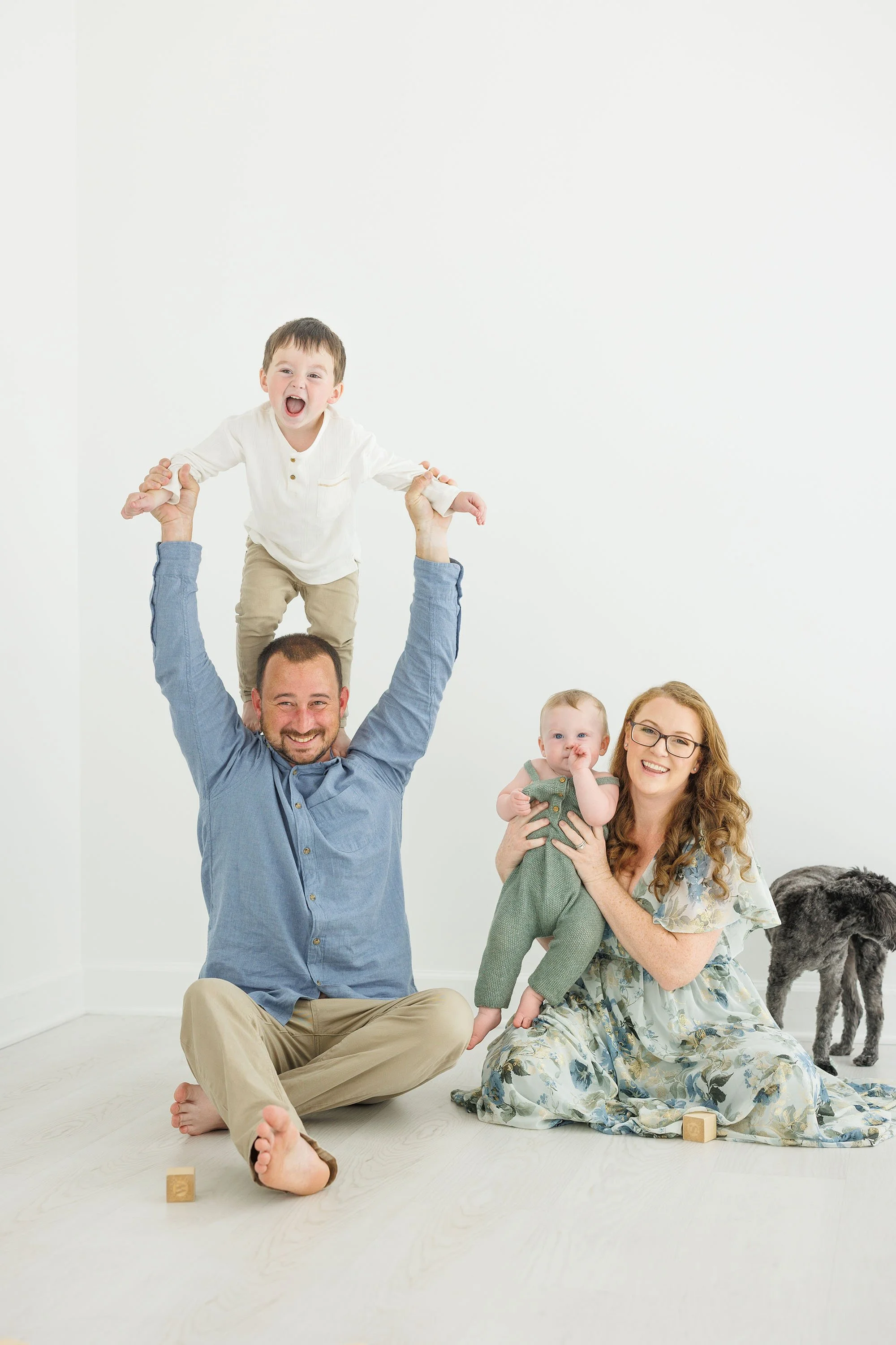 A family of four with two children, one on a man's shoulders, posing happily in a bright white room. The father is seated on the floor, with a woman holding a young boy, and a dog standing nearby.