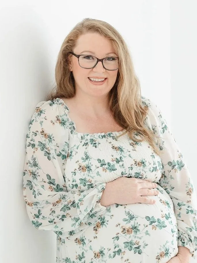 Pregnant woman with long red hair and glasses smiling, wearing a white floral dress, standing against a plain white background.