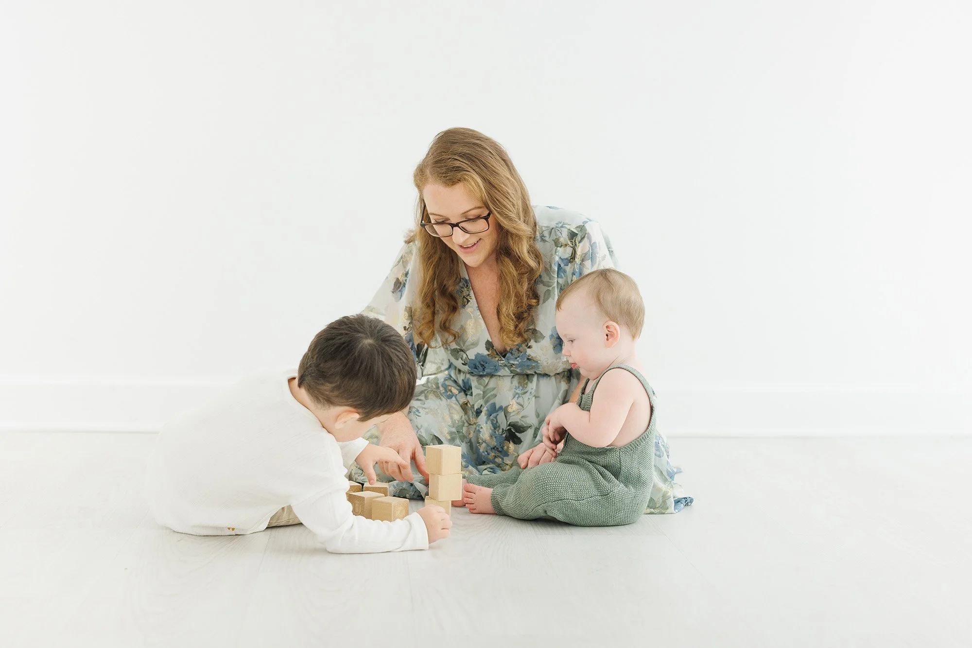 A woman with long hair and glasses playing with two children on a white floor against a plain white background.
