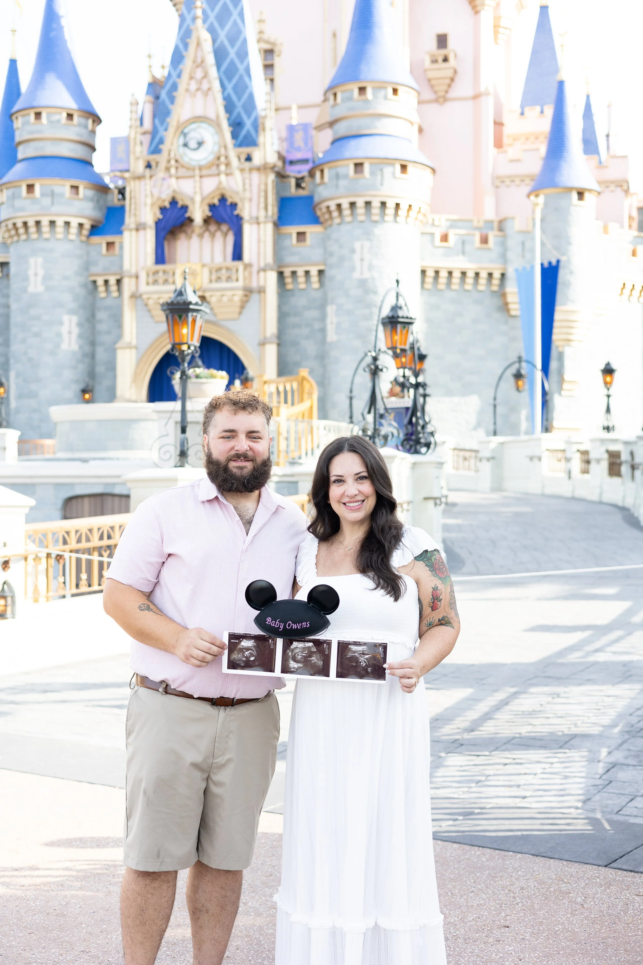A couple standing in front of Sleeping Beauty's castle at Disneyland, holding ultrasound photos and a Mickey Mouse hat that says 'Baby Owens', celebrating expecting a baby.