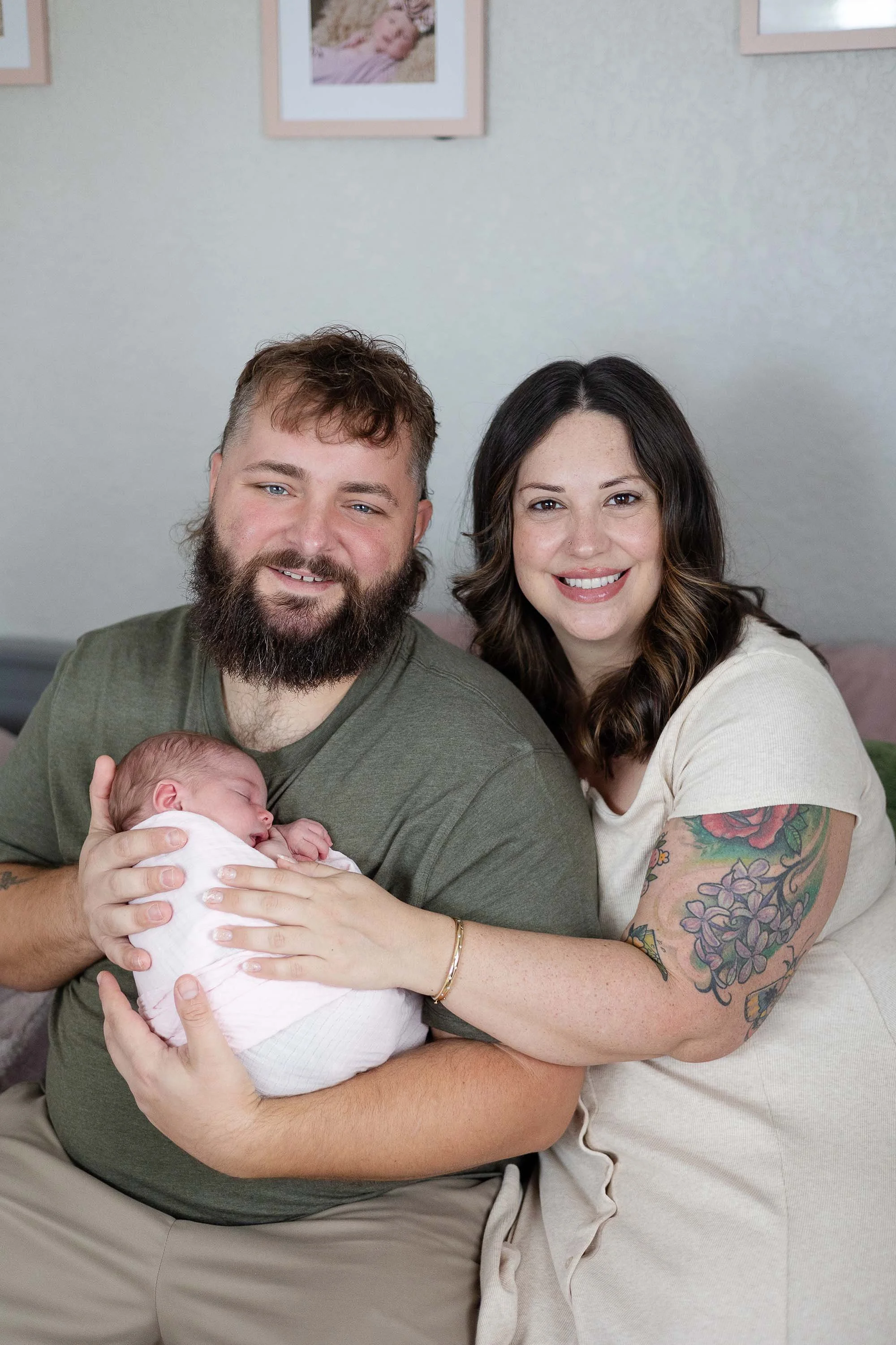 A family portrait of a man with a beard, woman with tattoos, and a newborn baby. The man is holding the baby, and the woman is smiling next to them. The background features framed pictures on the wall.