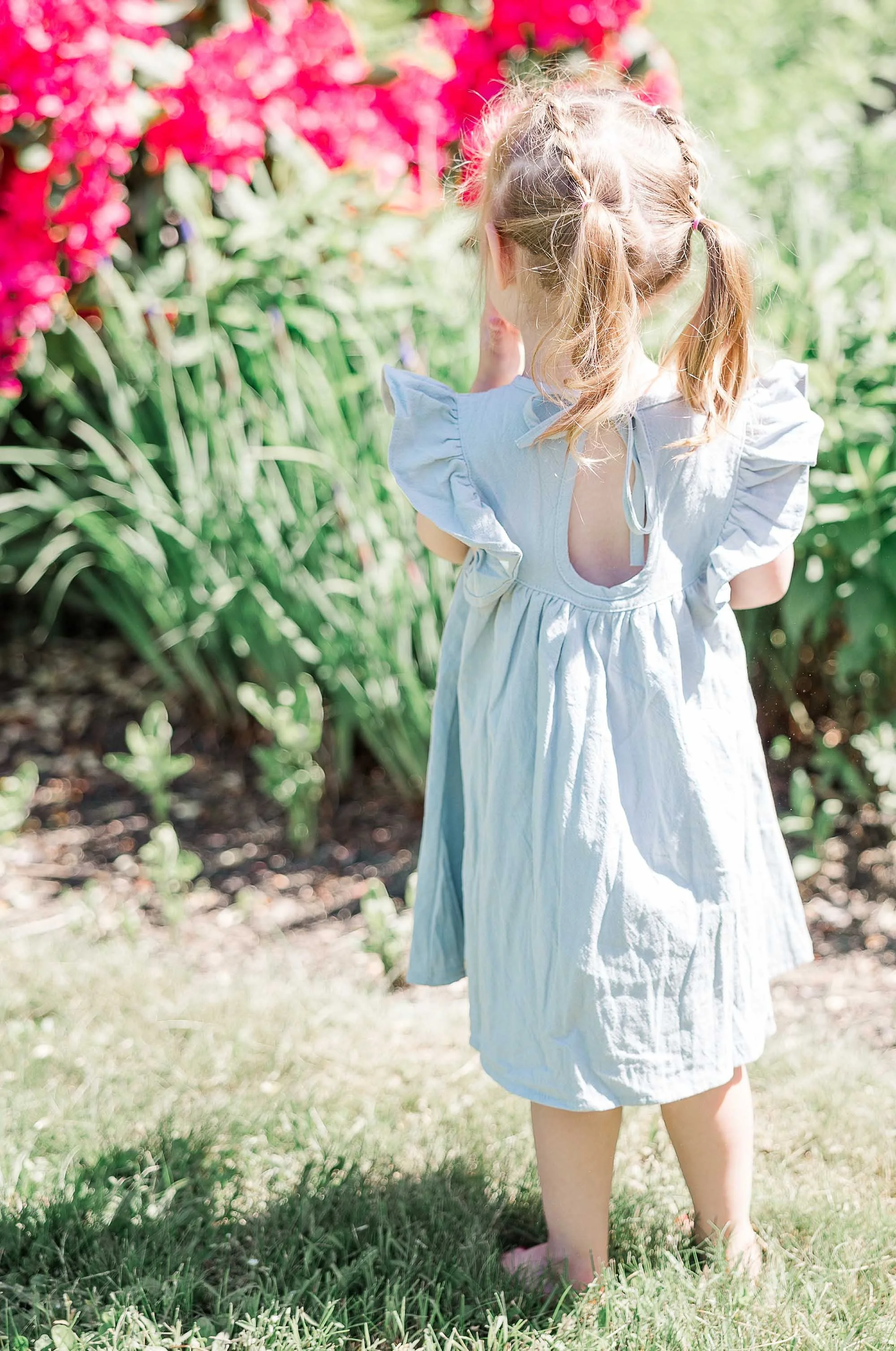 A young girl with blonde hair tied in pigtails, wearing a light blue dress with ruffled sleeves, is standing outdoors in a garden or park, facing away from the camera, surrounded by lush green grass and colorful pink flowers.