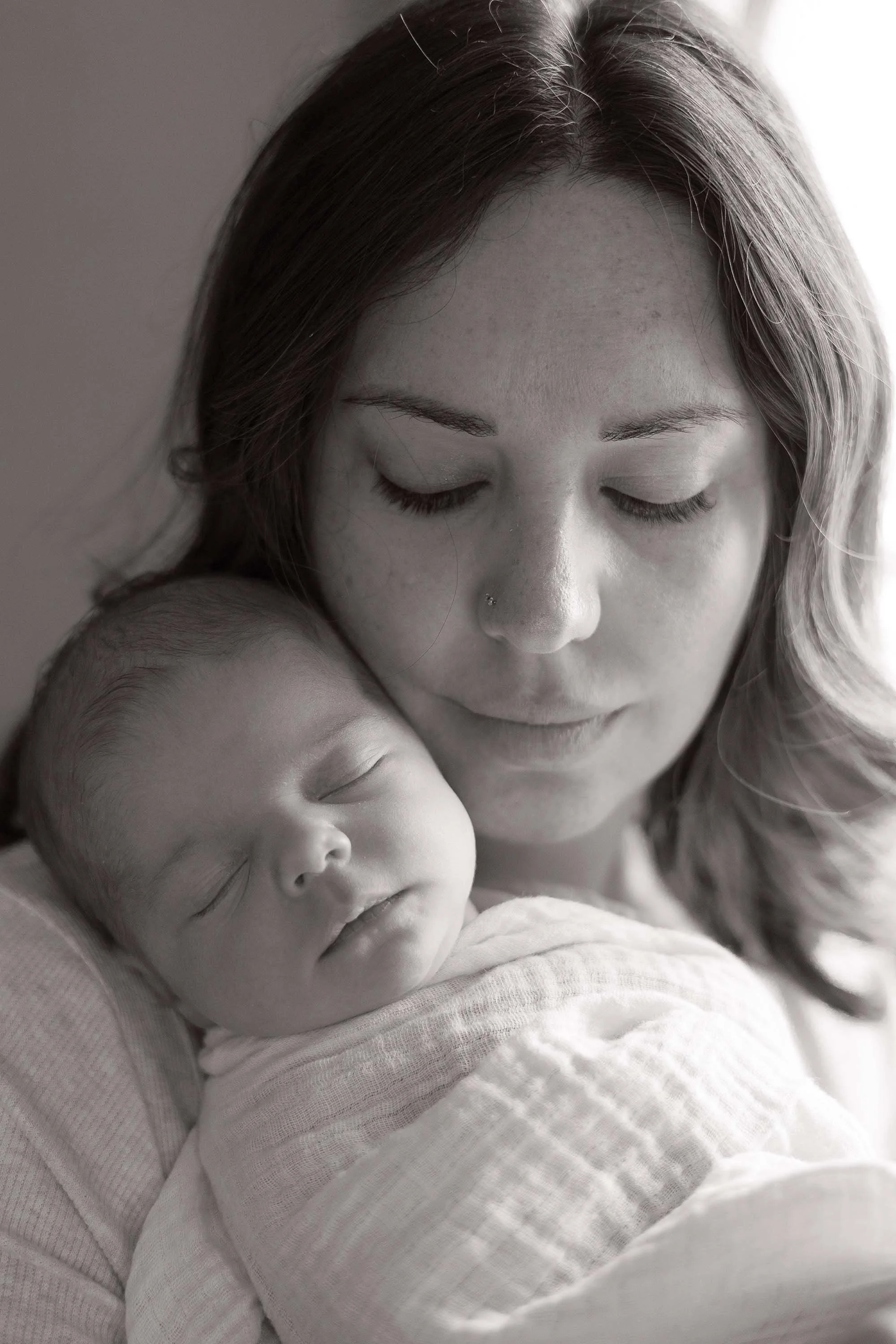 A woman with long hair, closed eyes, and a nose piercing gently holds a sleeping baby wrapped in a soft cloth.
