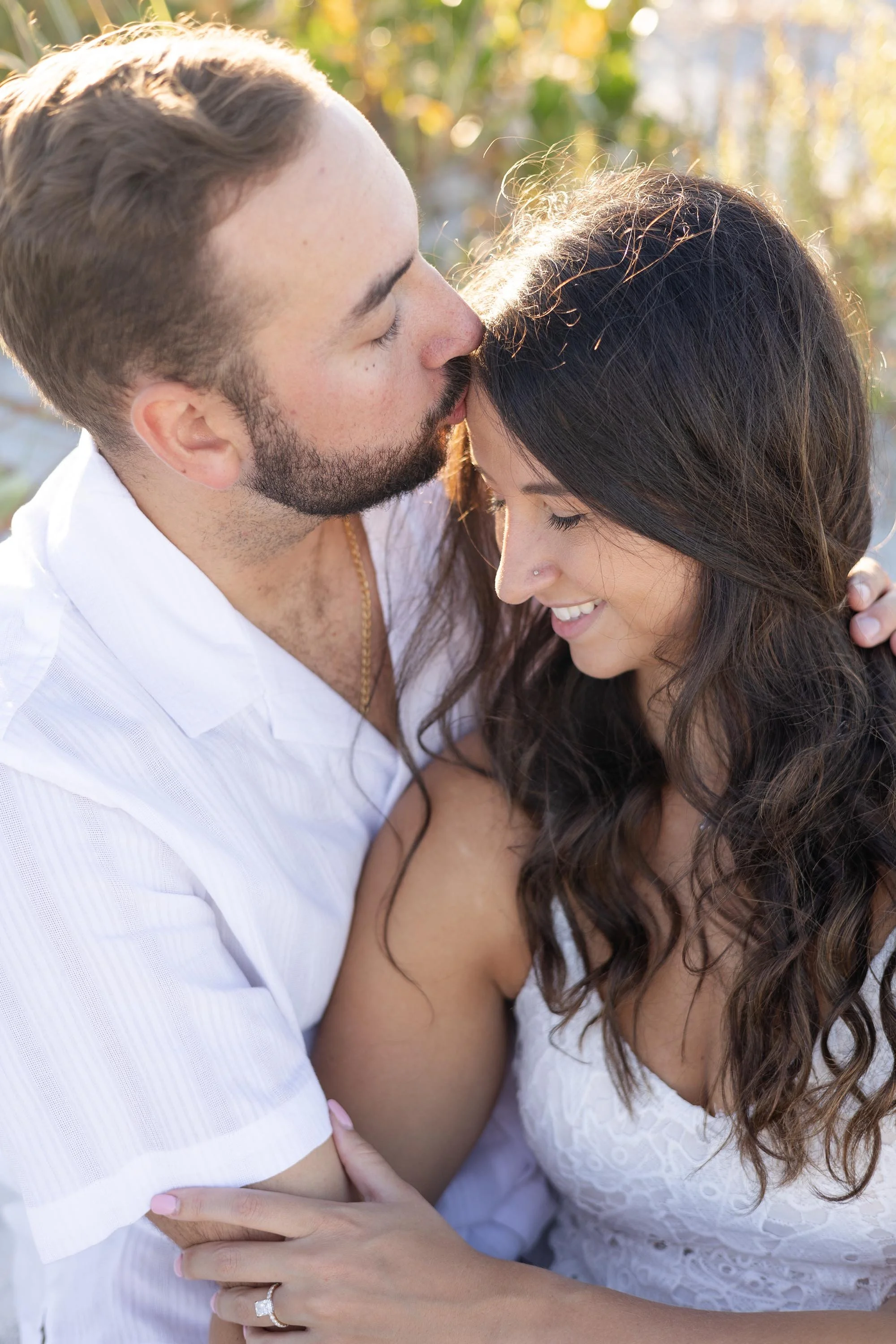 A man and woman embracing outdoors. The man is kissing the woman's forehead. Both are smiling and appear happy. The woman has long dark hair and a nose piercing. They are dressed in white and the background is blurred with trees and sunlight.