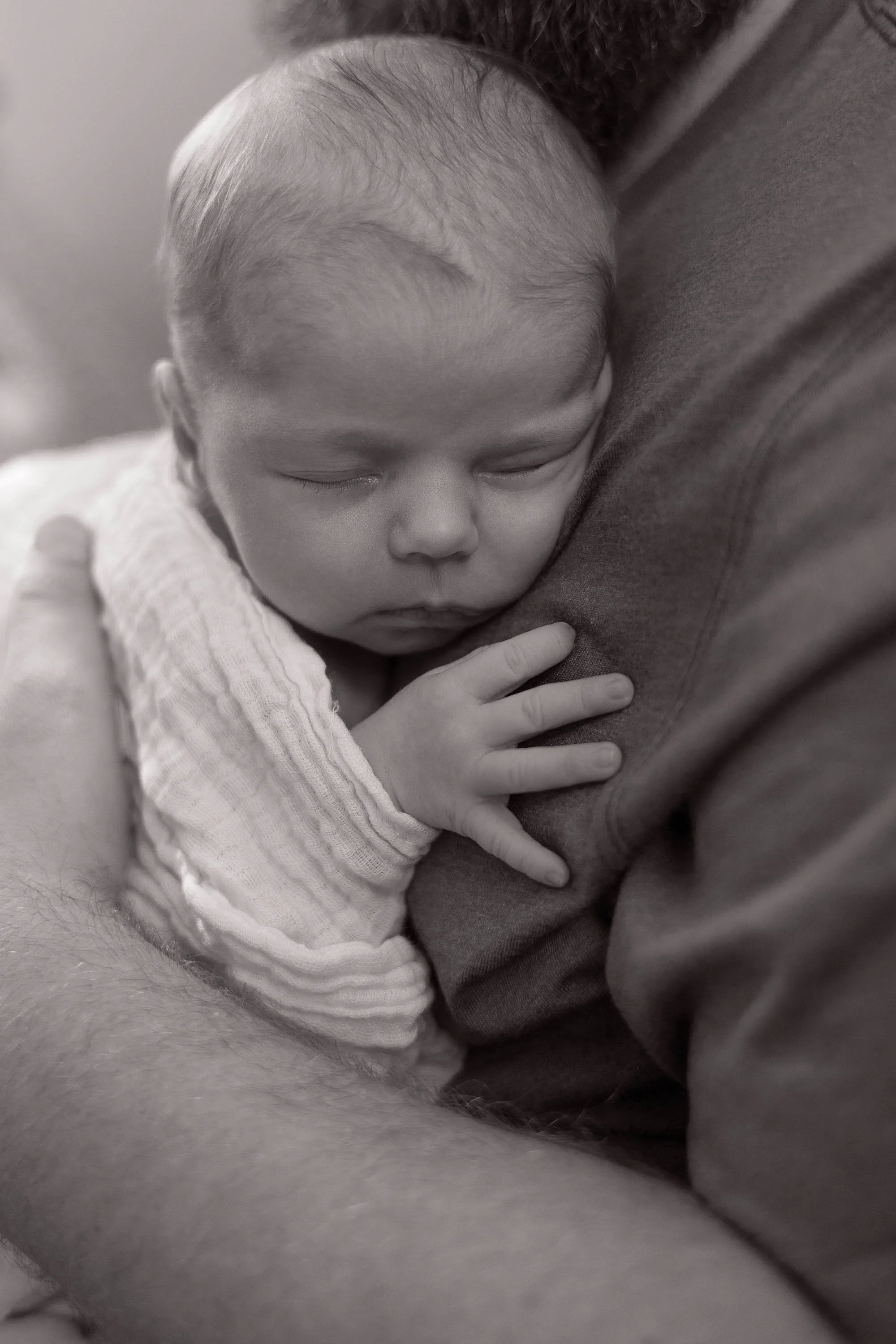 A sleeping baby with closed eyes, resting on an adult's chest, with one hand gently placed on the adult's arm. The baby is wearing a light-colored, long-sleeved outfit.