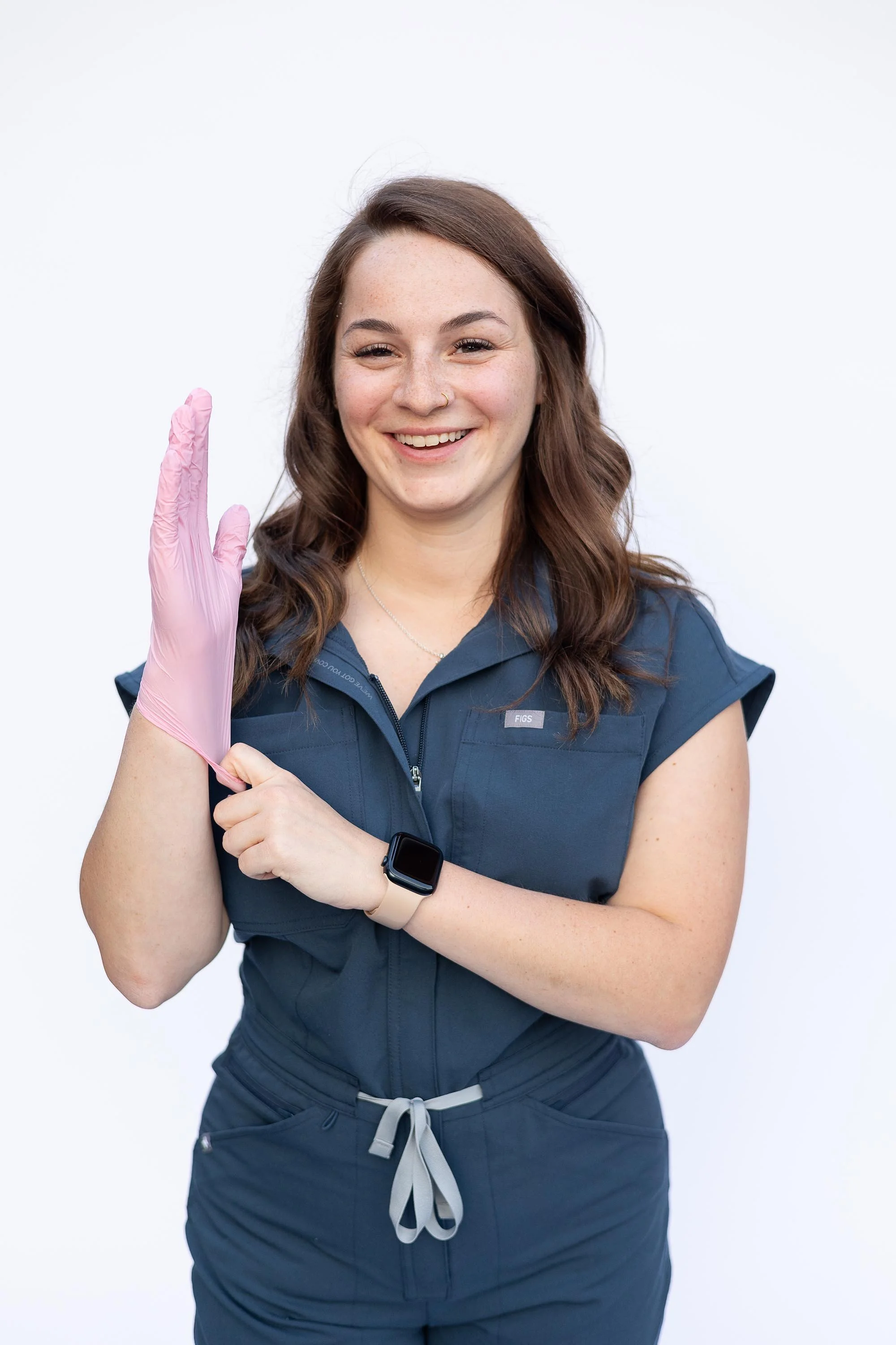 A woman in a navy blue uniform with a fitness tracker on her wrist, smiling and raising her left hand with a pink gloved finger pointing upward, against a plain white background.