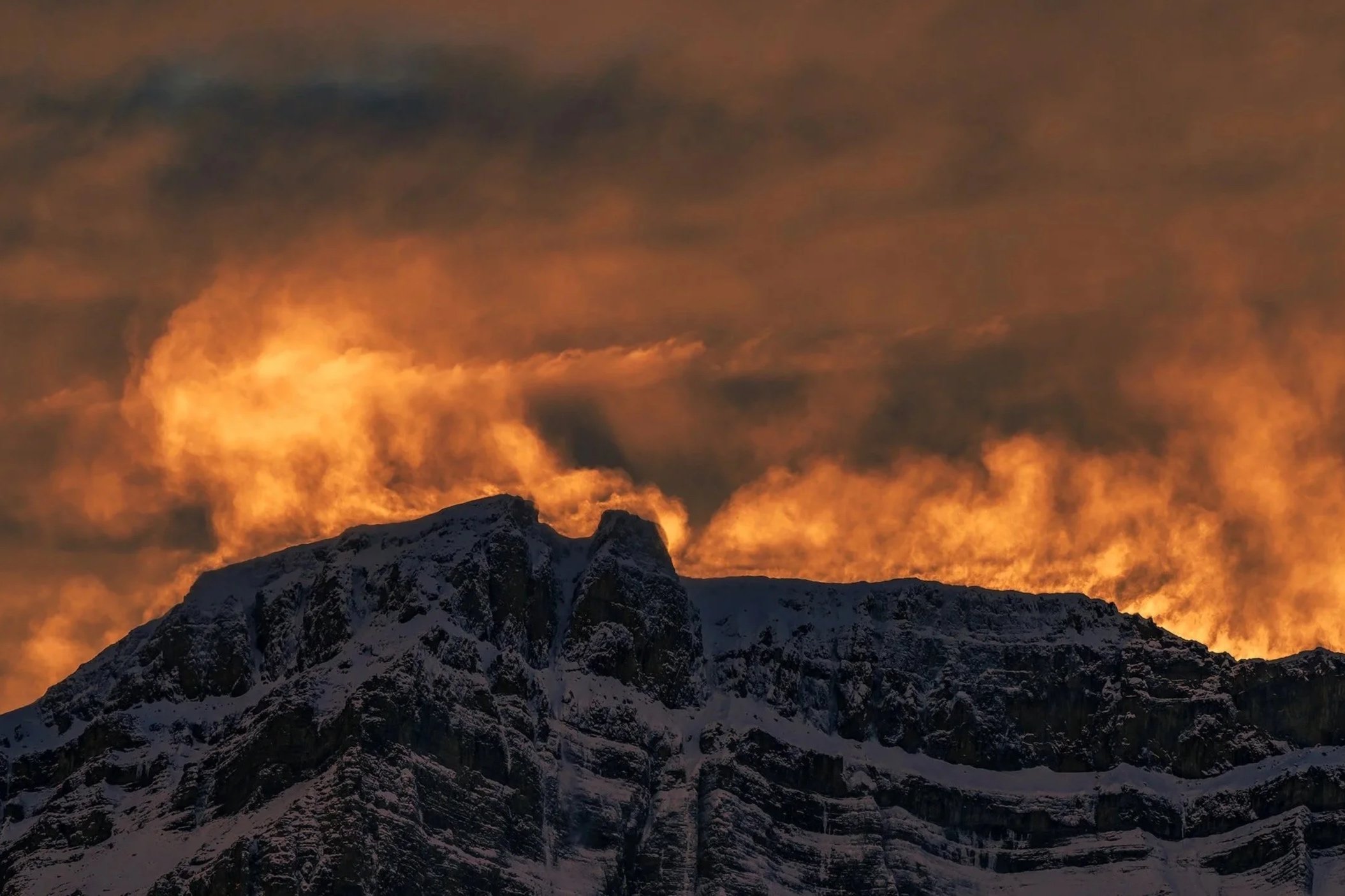 Snow-capped mountain peaks at sunset with orange and dark gray clouds in the sky.