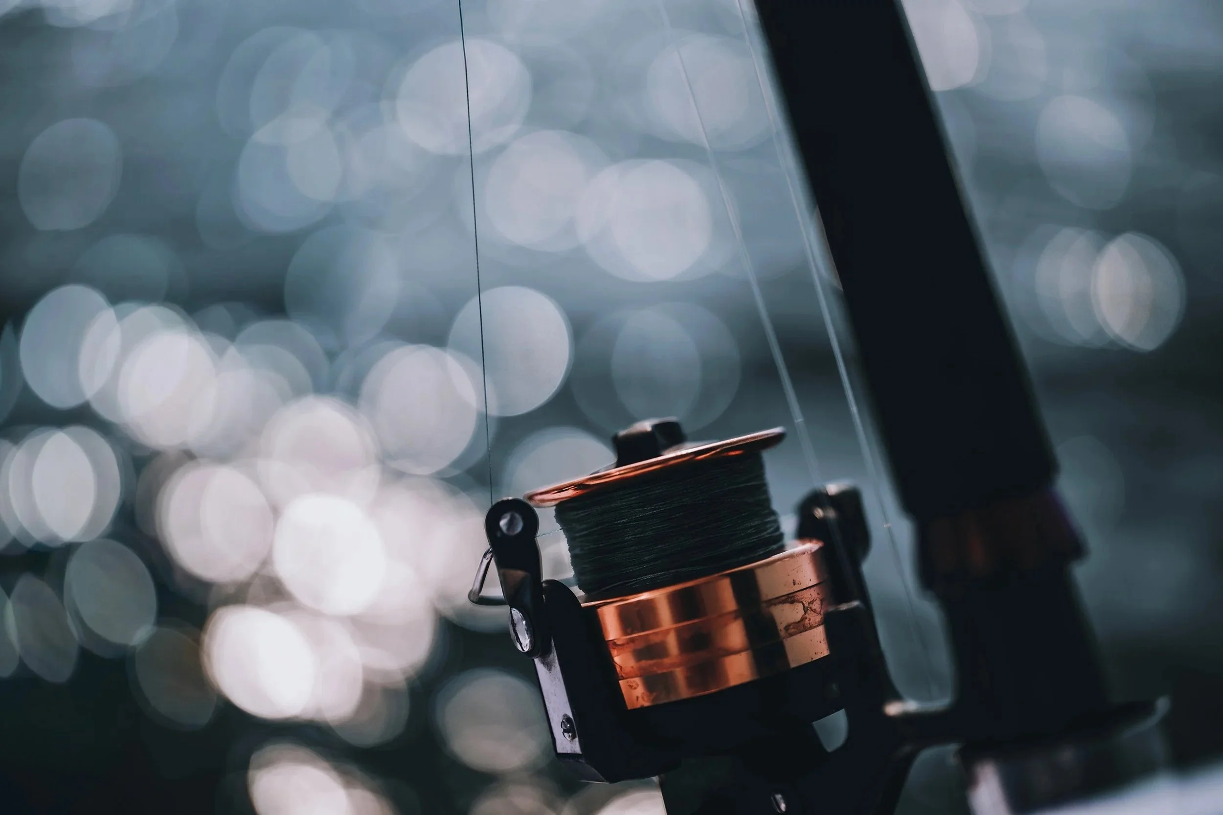Close-up of a fishing reel with a silver and copper finish, attached to a fishing rod, with blurred bokeh lights in the background.