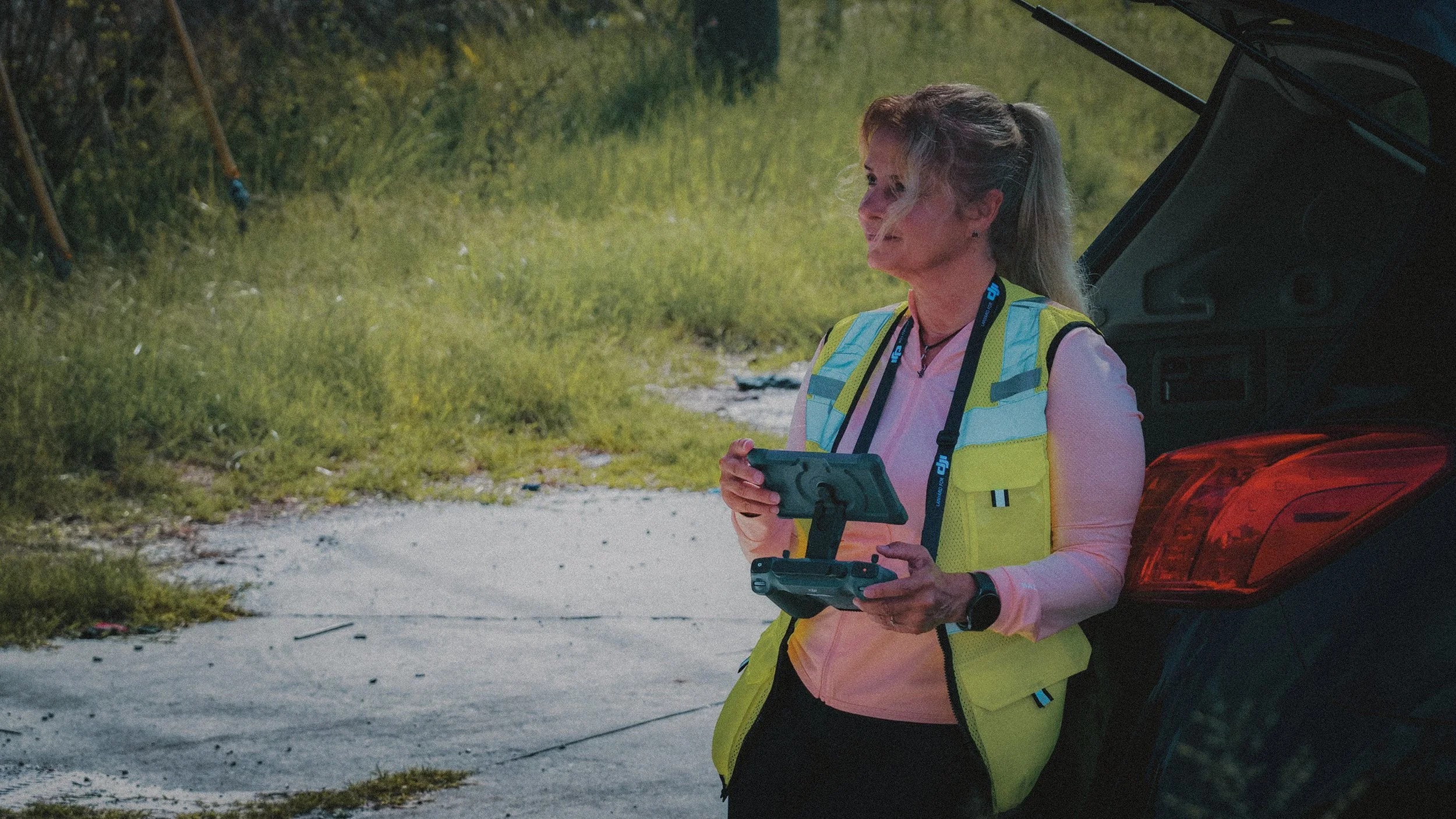 A woman standing behind a black car, holding a remote control, smiling, with her hair tied back, wearing a yellow safety vest and pink shirt, outdoors near a grassy area.