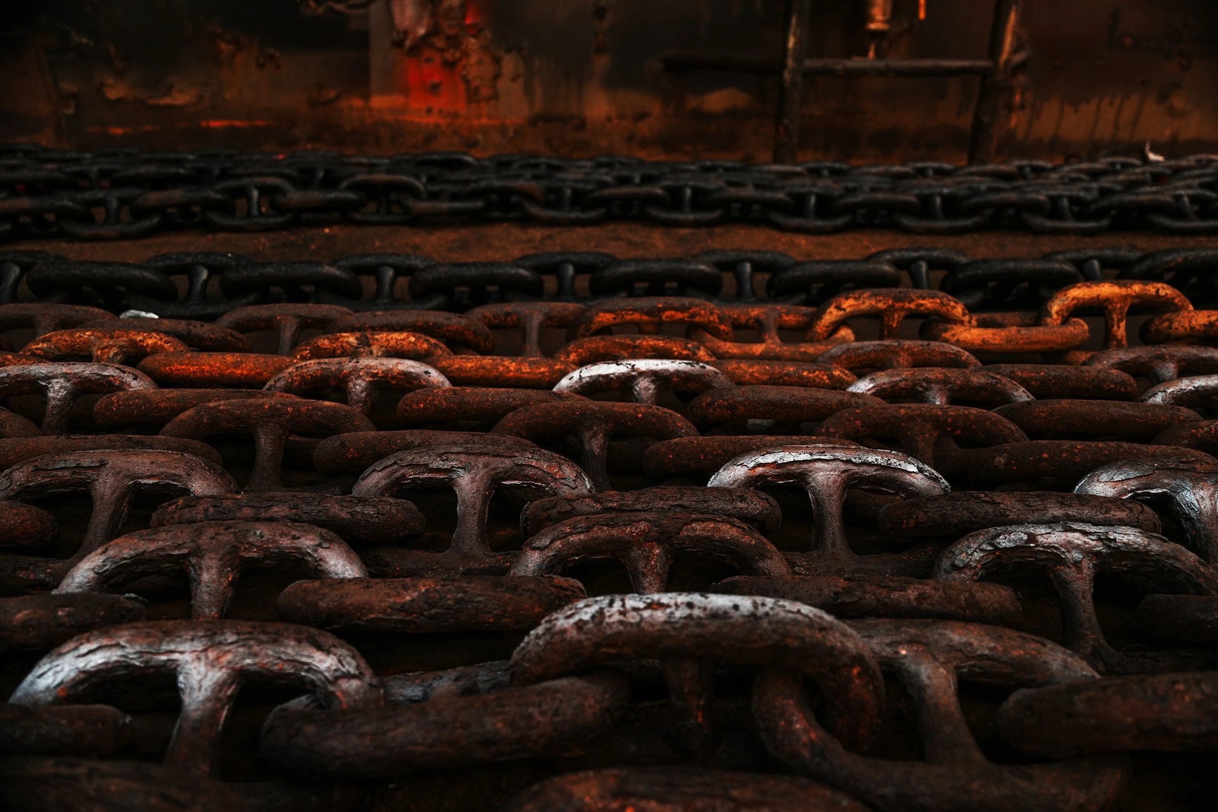 Close-up of rusty, black metal chains arranged on a surface, with a blurred background of rusty, red-brown metal surfaces.