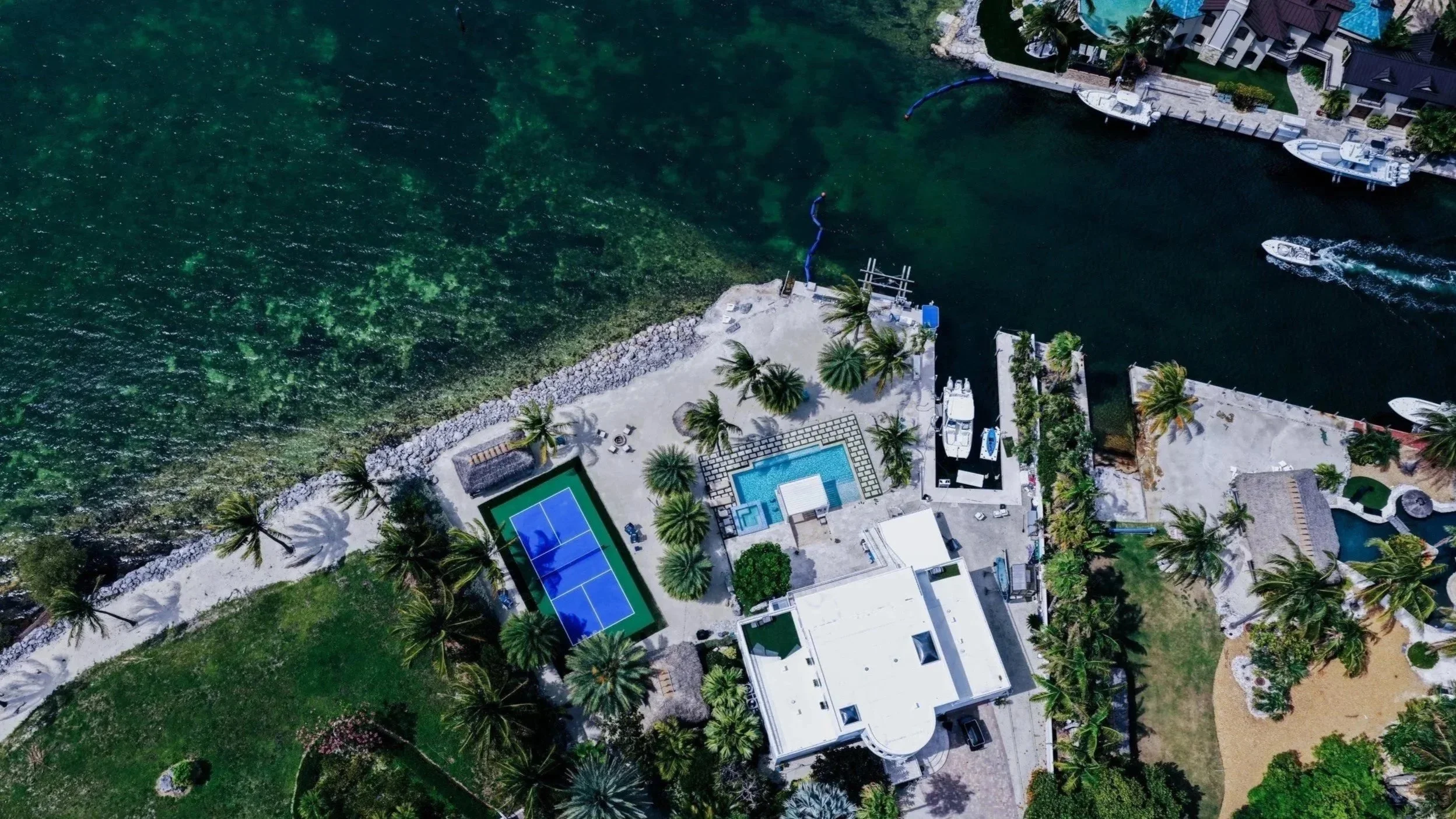 An aerial view of a beachfront property with swimming pool, tennis court, palm trees, boats docked along the water, and a water park slide extending into the water.