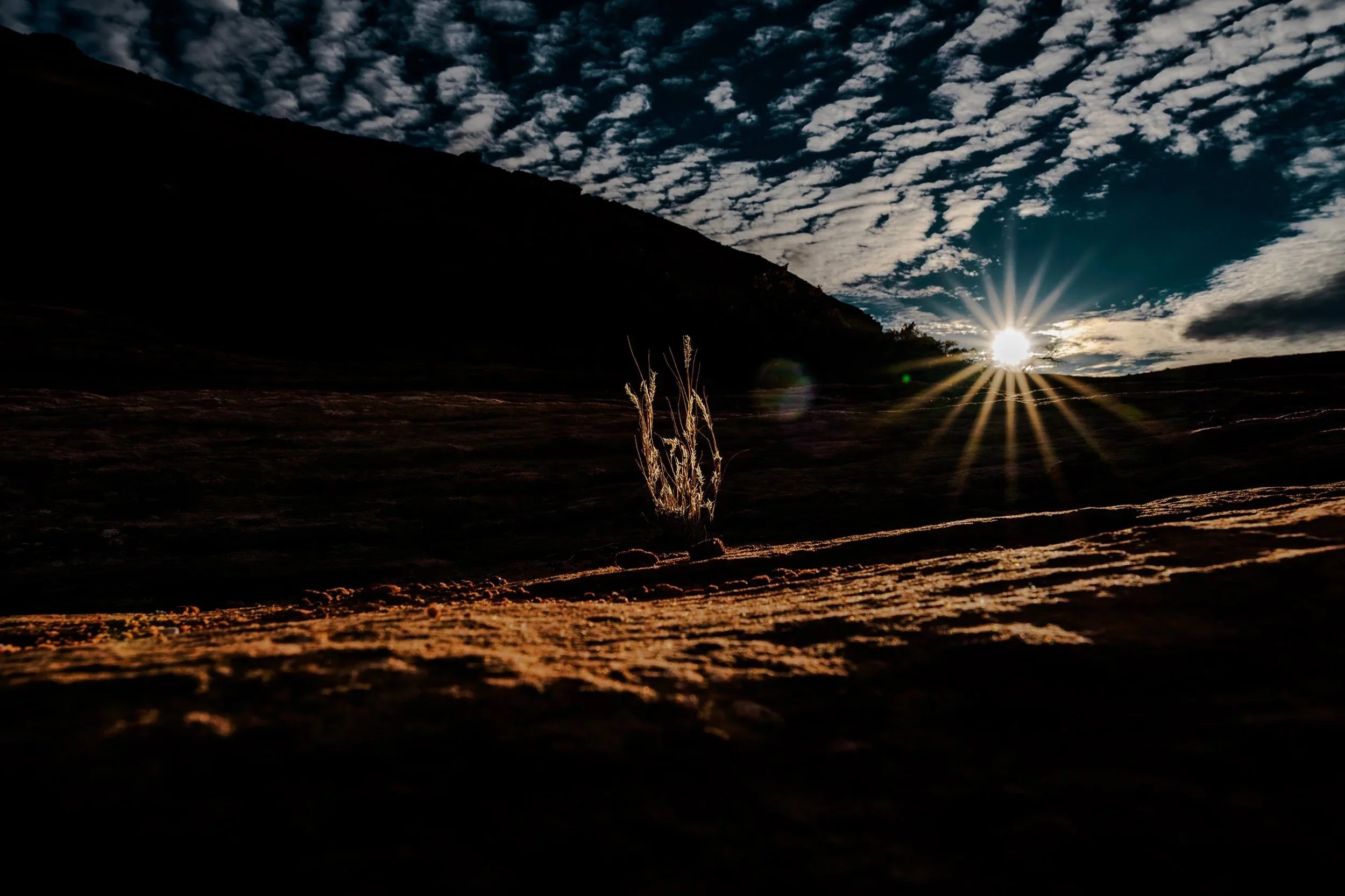 Sun setting behind hills with a dramatic sky filled with scattered clouds and rays of sunlight shining through, casting light on the sandy terrain in the foreground.