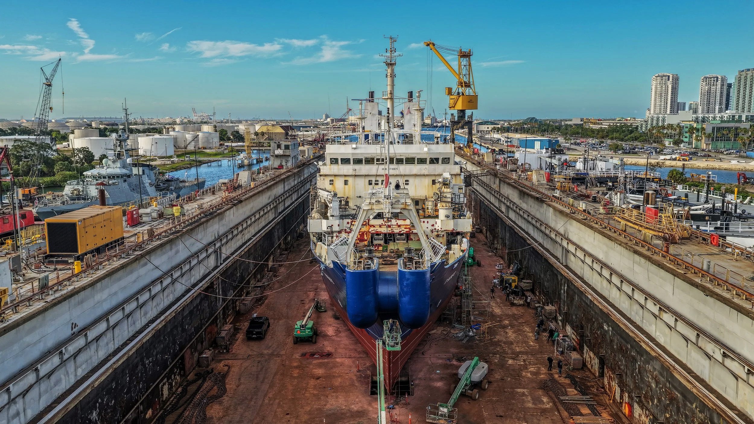 View of a shipyard with a large ship under construction inside a dry dock, surrounded by cranes, work areas, and nearby boats, with city buildings in the background under a blue sky.
