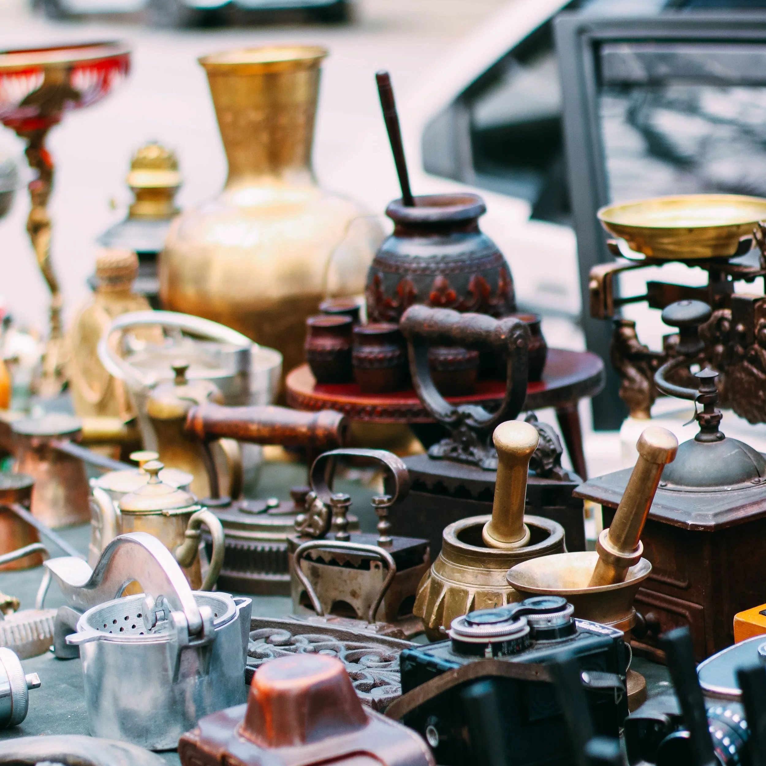Various metal and ceramic objects, including vases, bowls, and a vintage lamp, displayed on tables at an outdoor market.