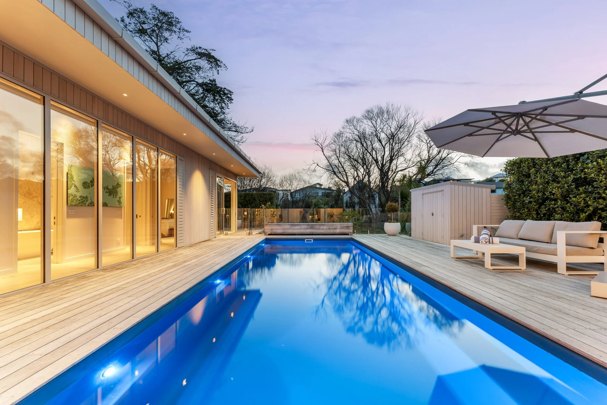 An outdoor backyard with a rectangular swimming pool, surrounding wooden deck, in Matakana Village, Auckland, New Zealand. The house has large glass doors and windows, and there are trees and a fence in the background during dusk.