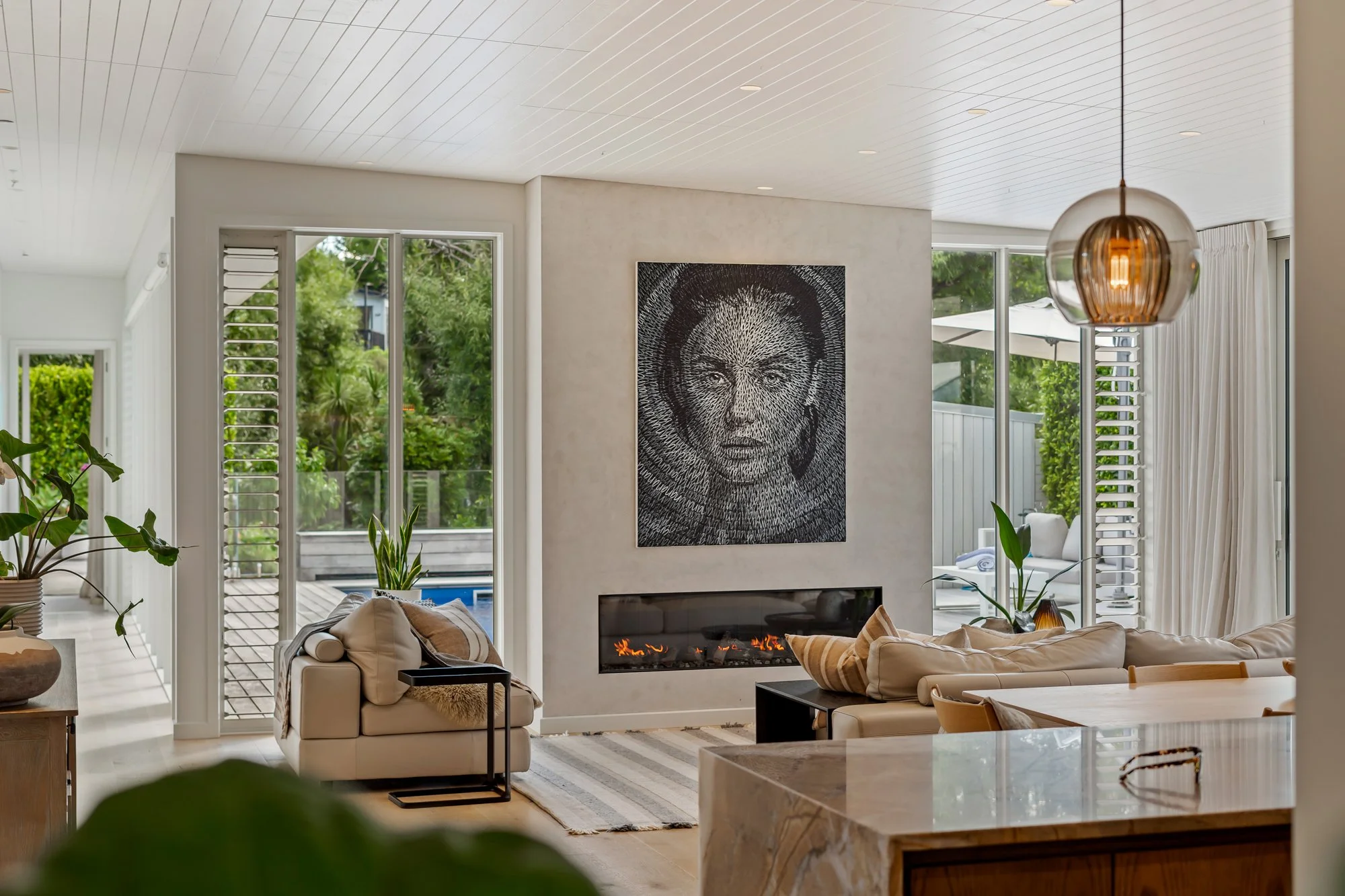A premium home in Matakana Village, Auckland, New Zealand, with modern living room with a fireplace, large windows, and artwork of a woman's face on the wall.