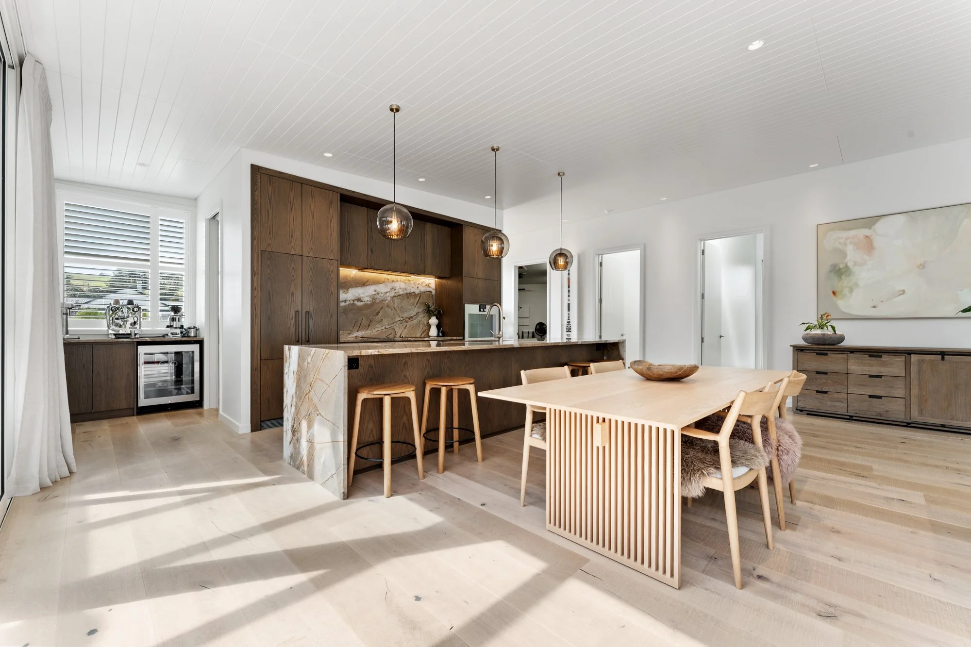 A premium home in Matakana Village, Auckland, New Zealand, with modern kitchen and dining area with light wood flooring, white walls, dark wood cabinetry, marble accents, and a light wooden dining table with chairs, pendant lighting, and a sideboard.