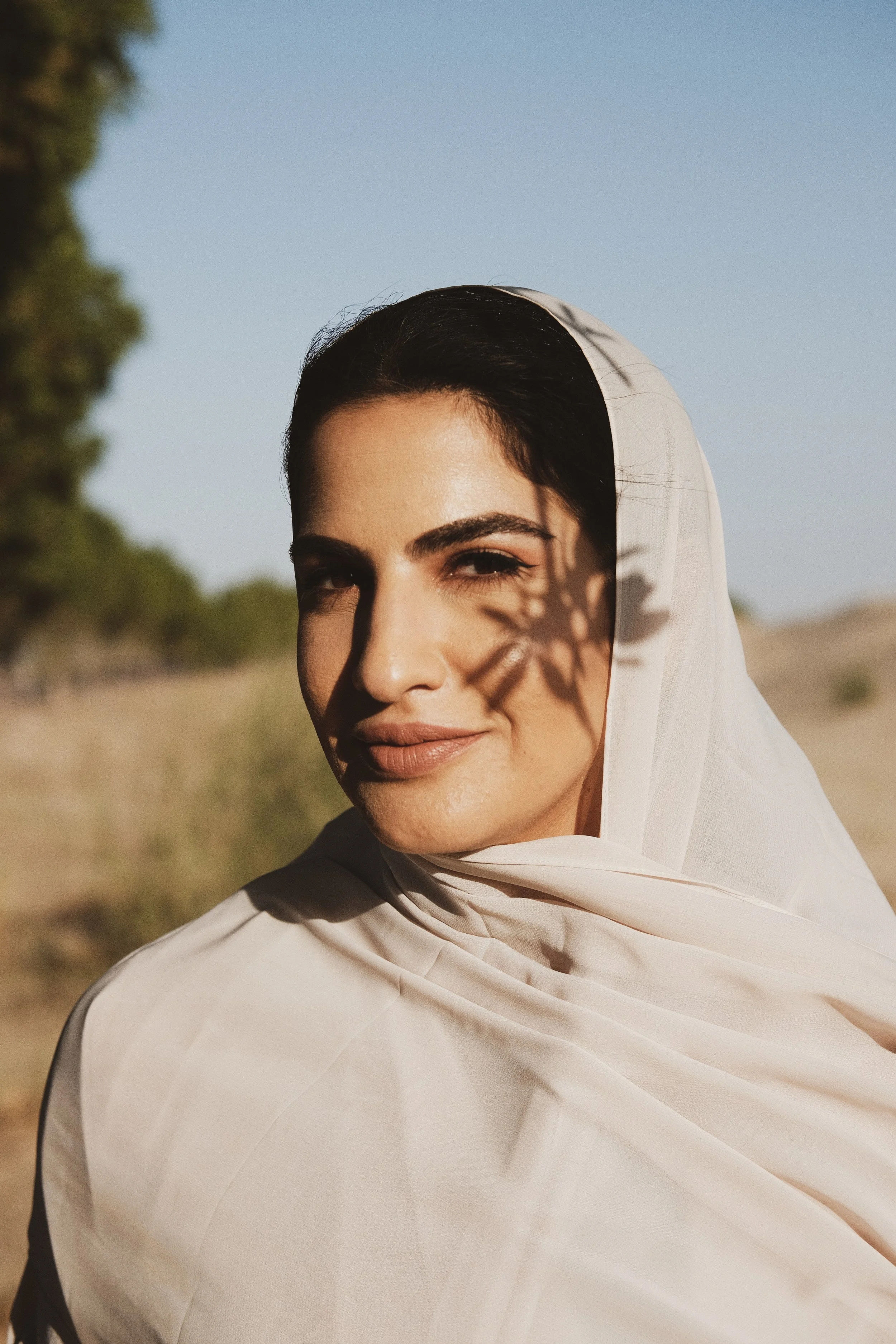 A woman with dark hair, partially covered with a white scarf, standing outdoors in sunny weather, with a shadow of a plant on her face, in a natural setting with trees and a clear blue sky.