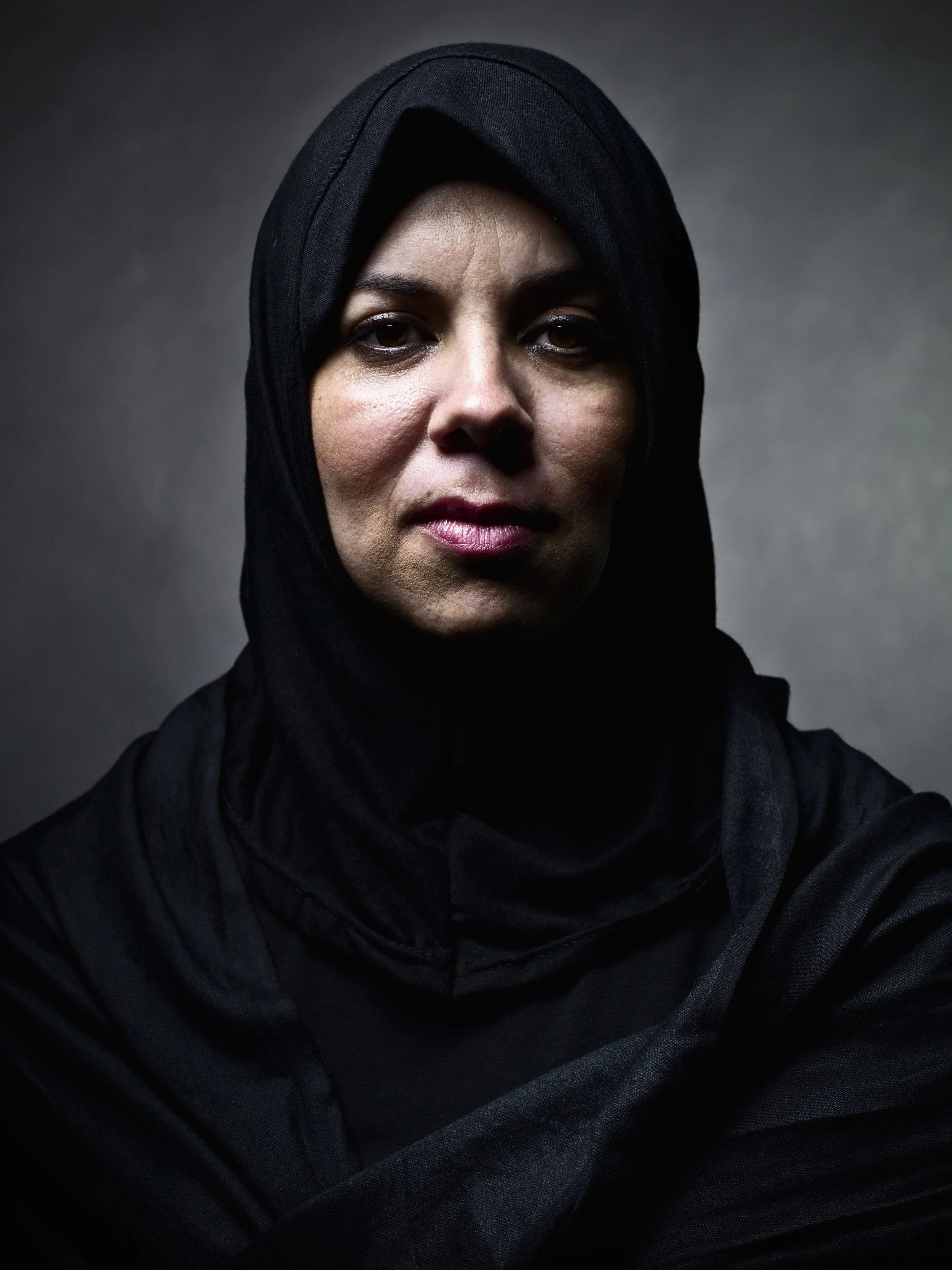 A woman wearing a black hijab and black clothing, looking directly at the camera with a serious expression, against a dark gray background.