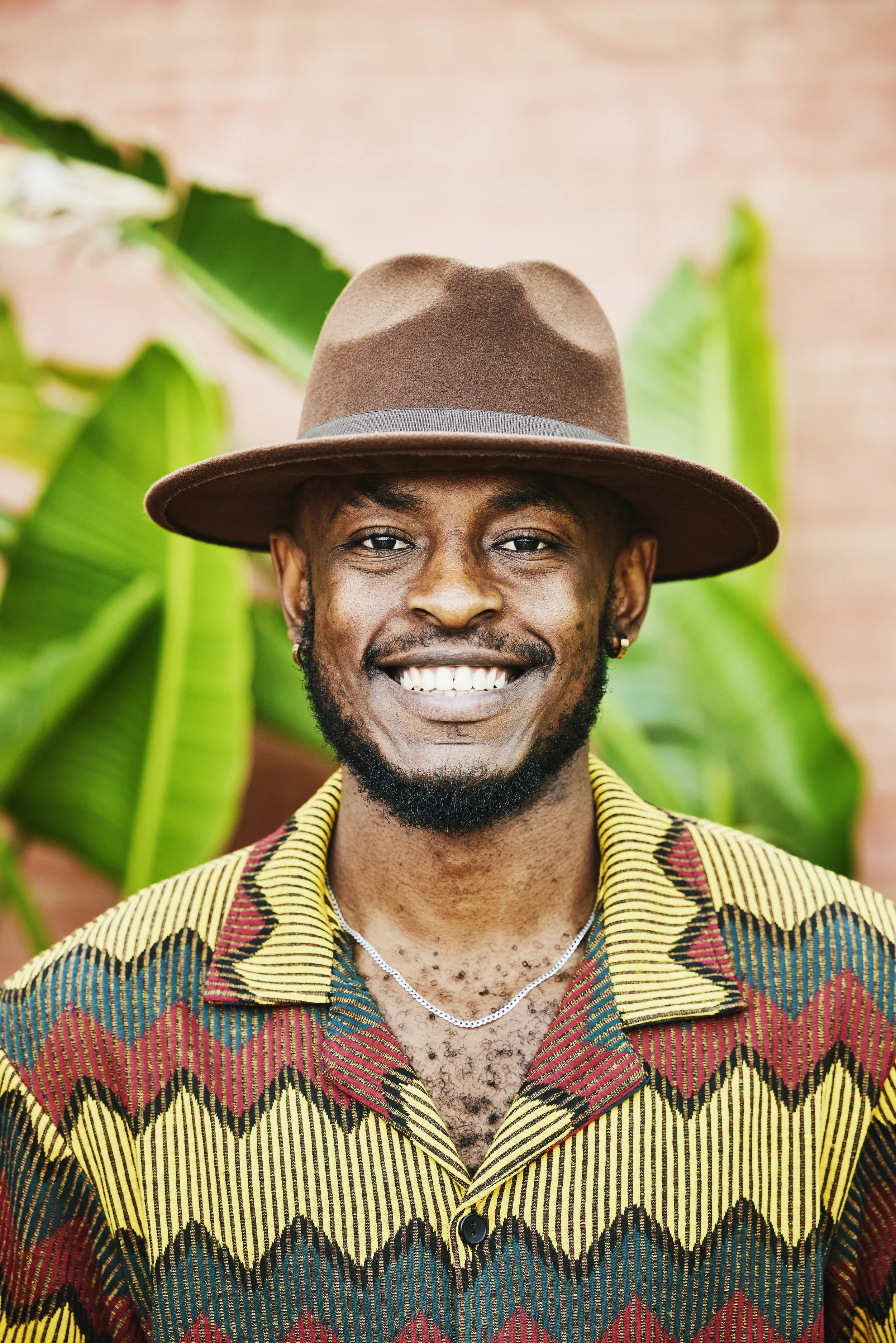 A smiling man wearing a wide-brimmed brown hat, a colorful patterned shirt, a silver chain, and earrings, standing in front of large green tropical leaves and a pink brick wall.
