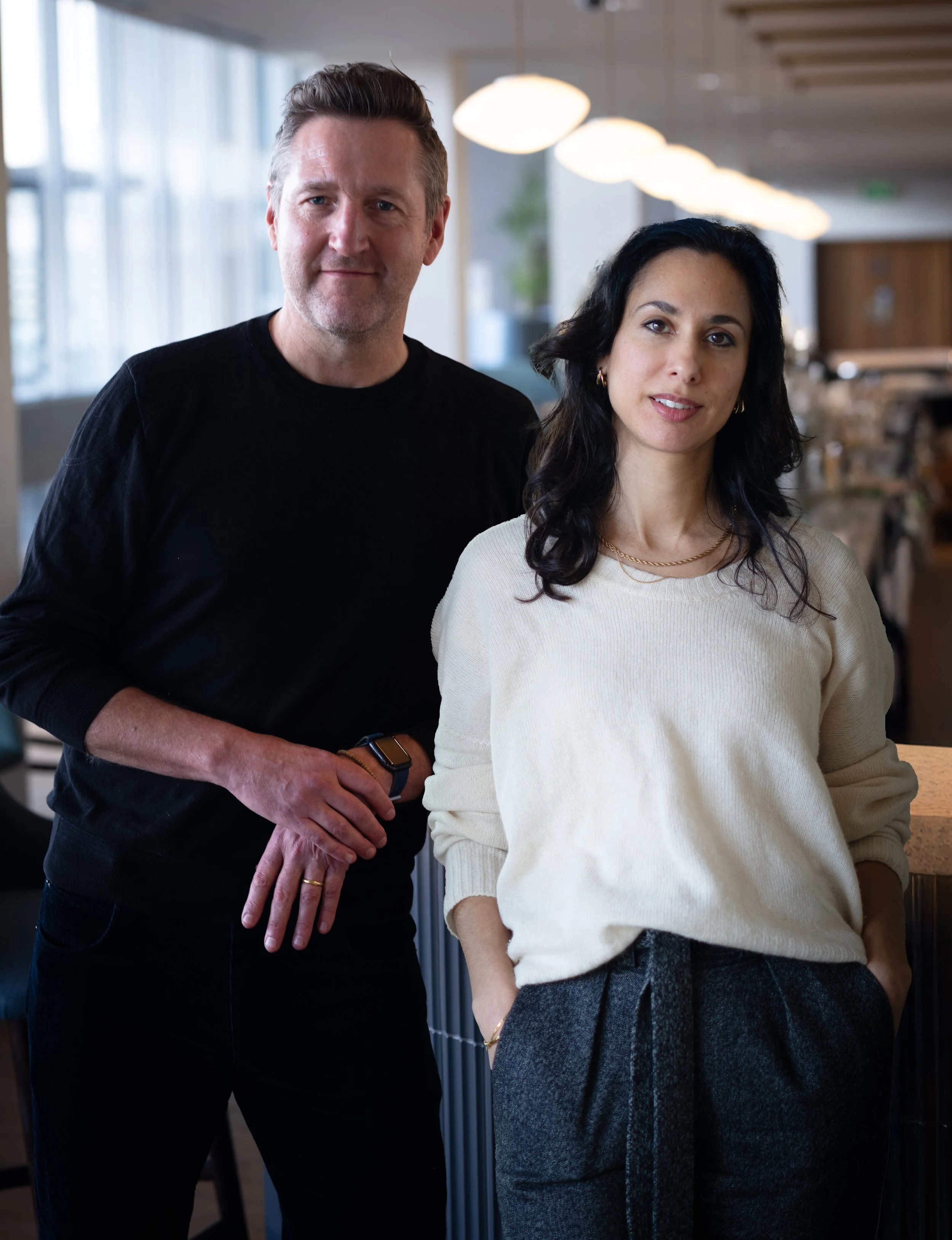 A man and woman standing in a modern, well-lit indoor space, with the man behind and to the left of the woman. The man has short gray hair, wears a black long-sleeve shirt, and a smartwatch. The woman has shoulder-length dark hair, wears a cream-colored sweater, gold jewelry, and gray pants. Both are looking at the camera.