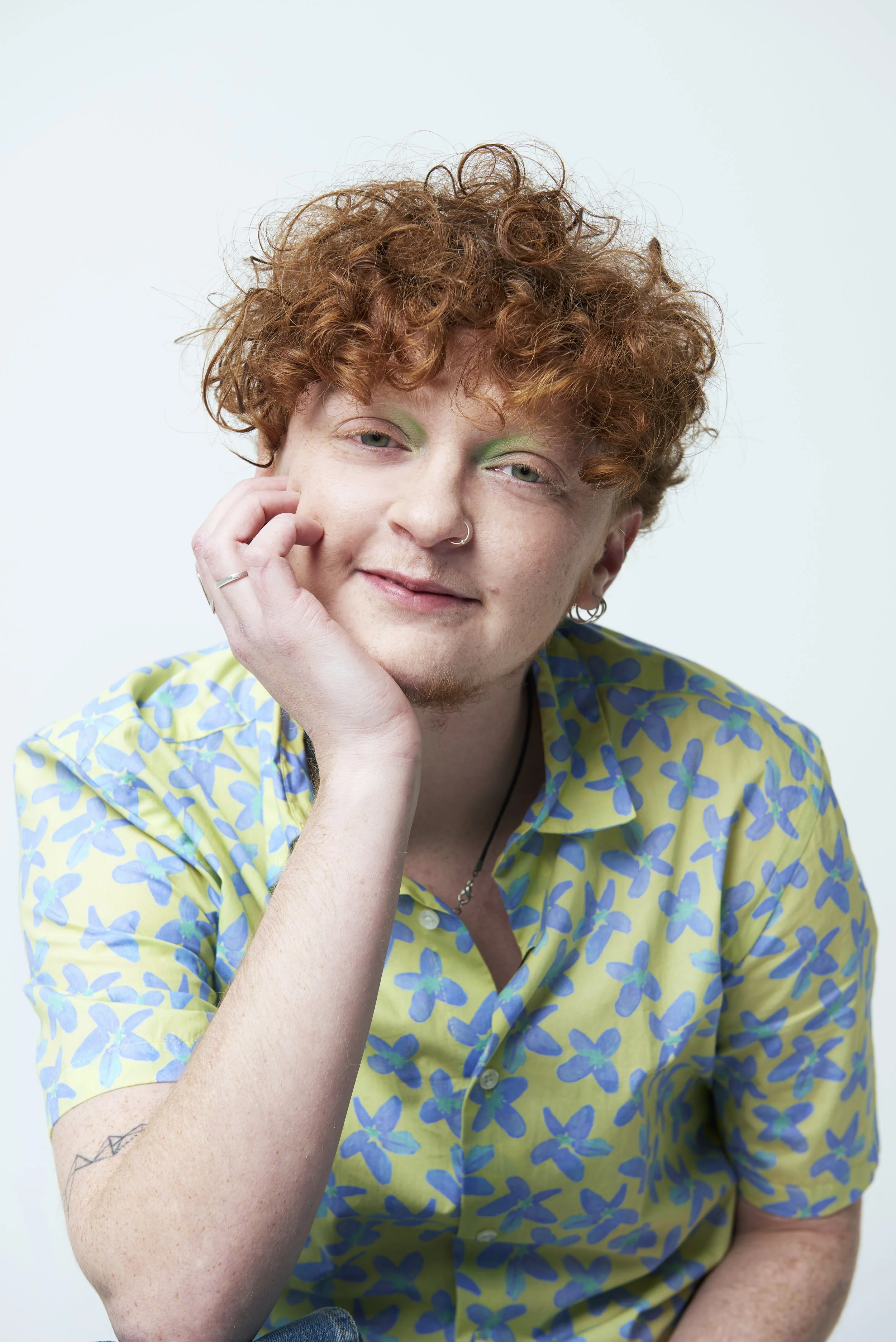 A person with curly red hair, wearing a yellow shirt with blue floral patterns, resting their chin on one hand, smiling softly.