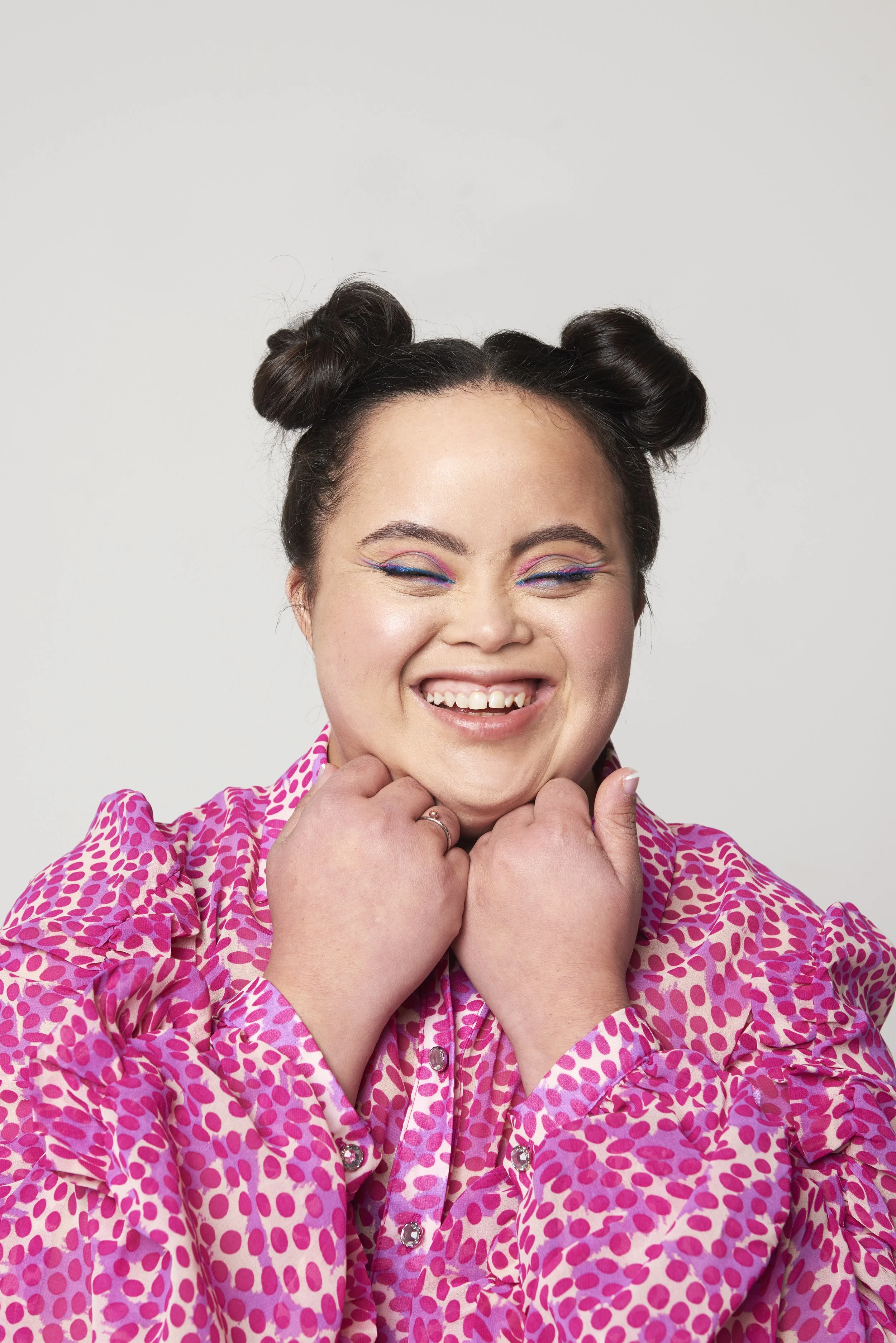 A woman with dark hair styled in two buns, wearing colorful makeup and a pink, patterned blouse, smiling and holding her fists near her chin.