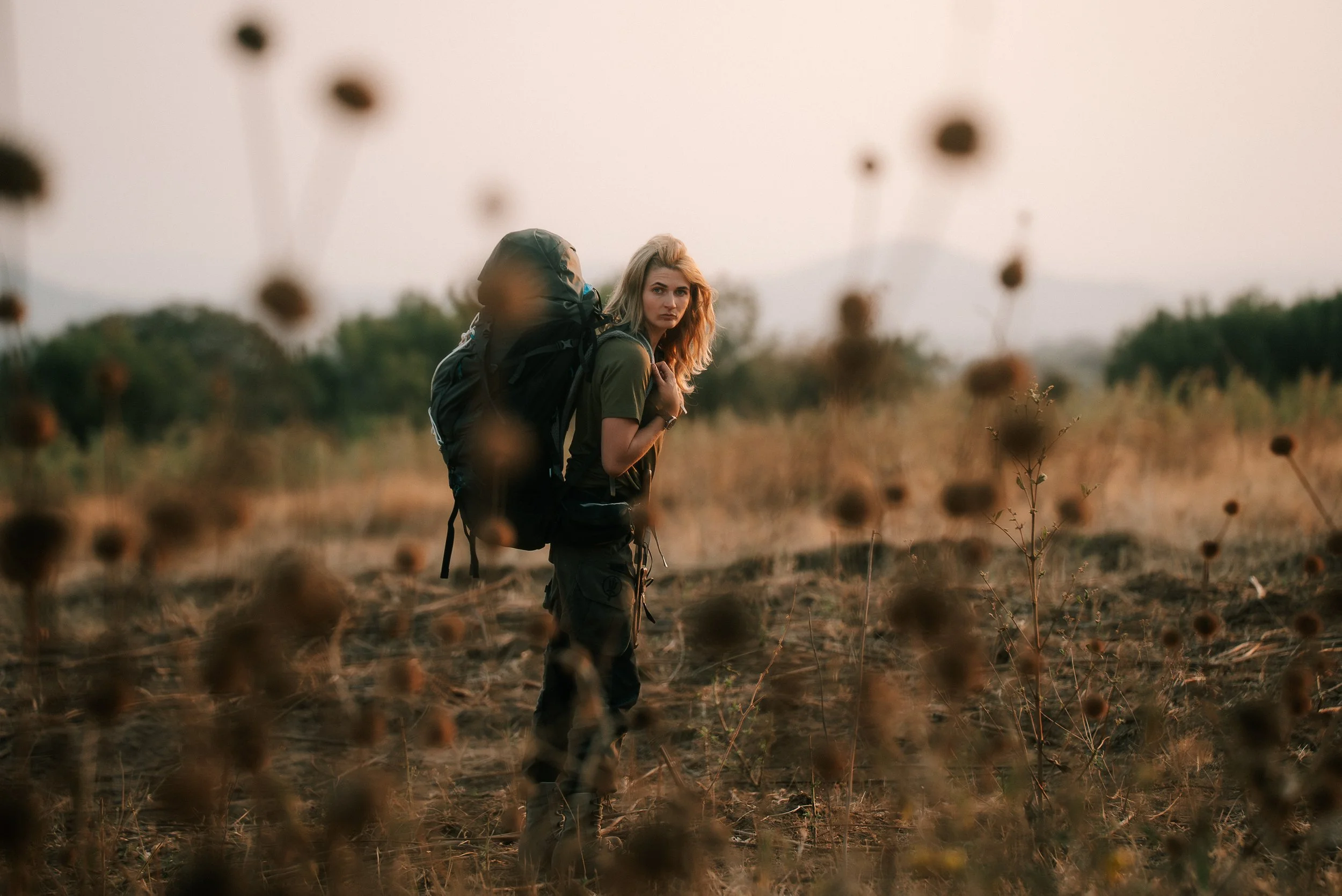 A woman with blonde hair wearing outdoor hiking gear, carrying a large backpack, standing in a field of dry plants at sunset, looking at the camera.