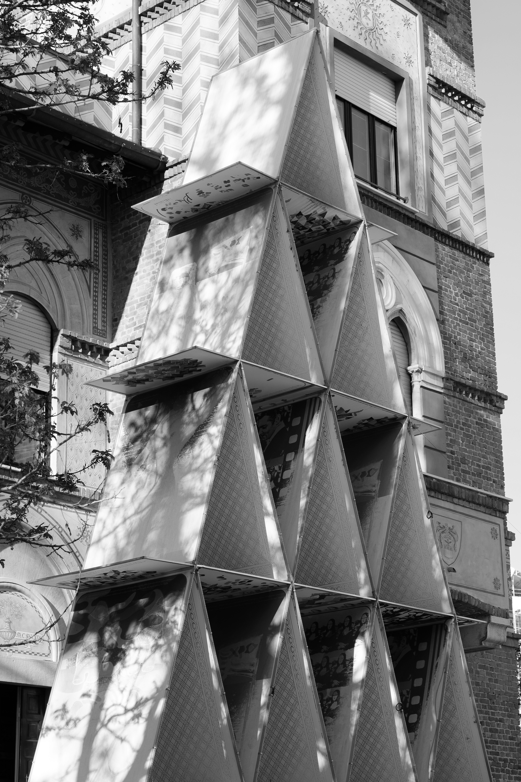 Edificio storico con facciata in mattoni e decorazioni, sulla quale si erge una struttura moderna composta da pannelli metallici disposti a scudo. Ombre di alberi sono visibili sulle superfici della struttura.