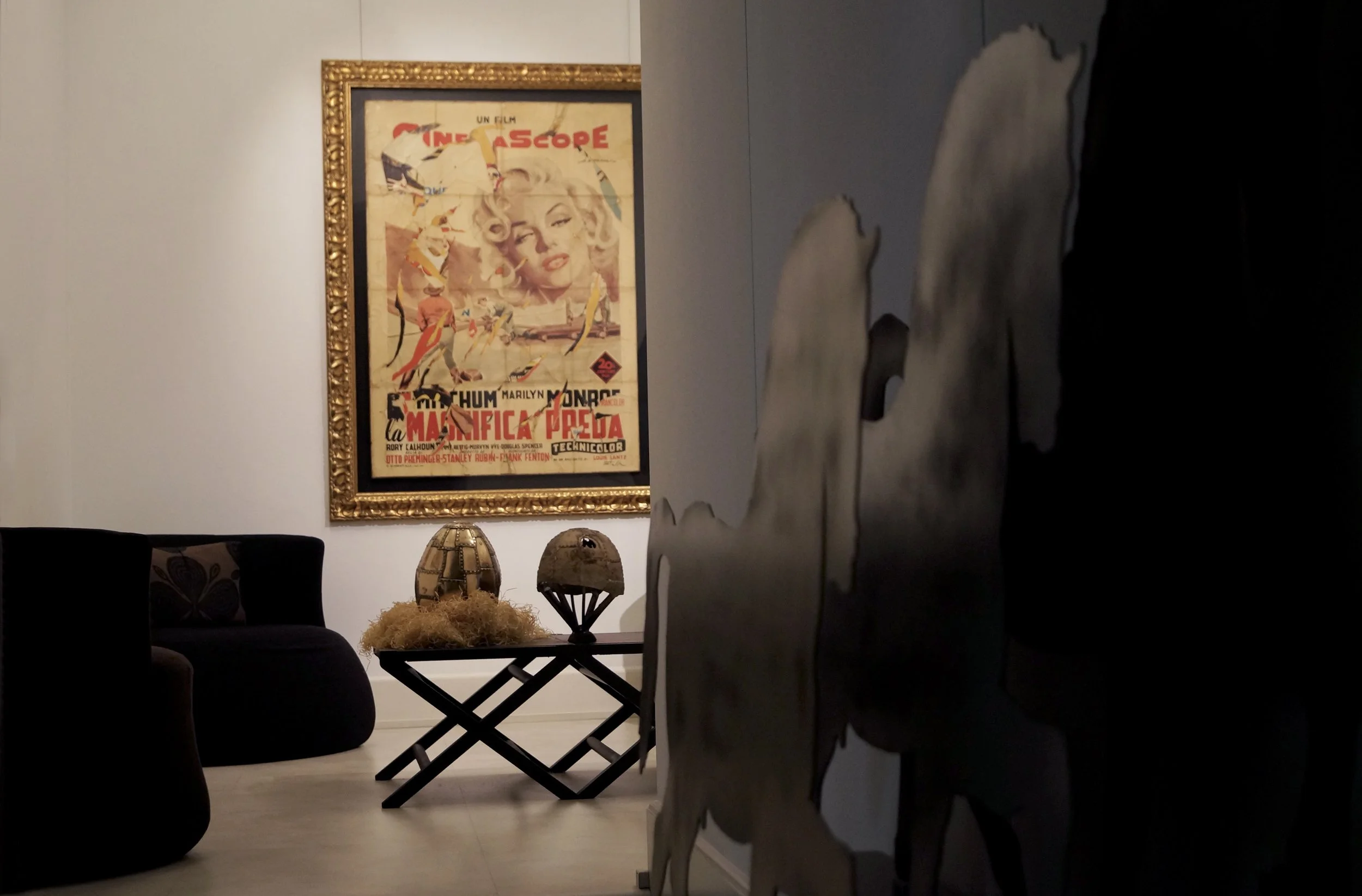 Interno di un ambiente con un quadro in cornice dorata sulla parete; parte di una sedia e tavolini decorativi in primo piano; pareti chiare e graziosi oggetti decorativi.