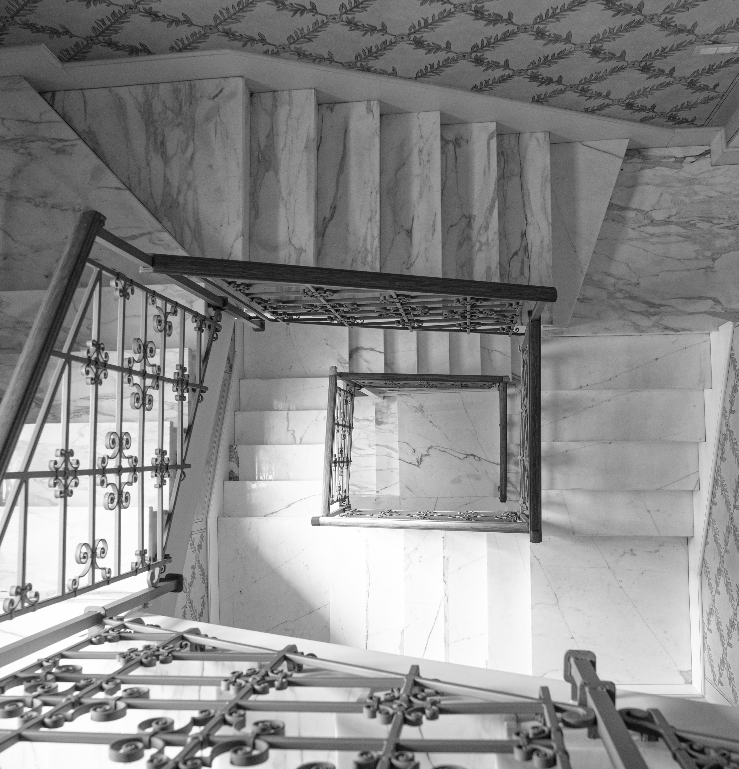 Immagine in bianco e nero di una scala di marmo con un ballatoio in ferro battuto decorato, vista dall'alto.