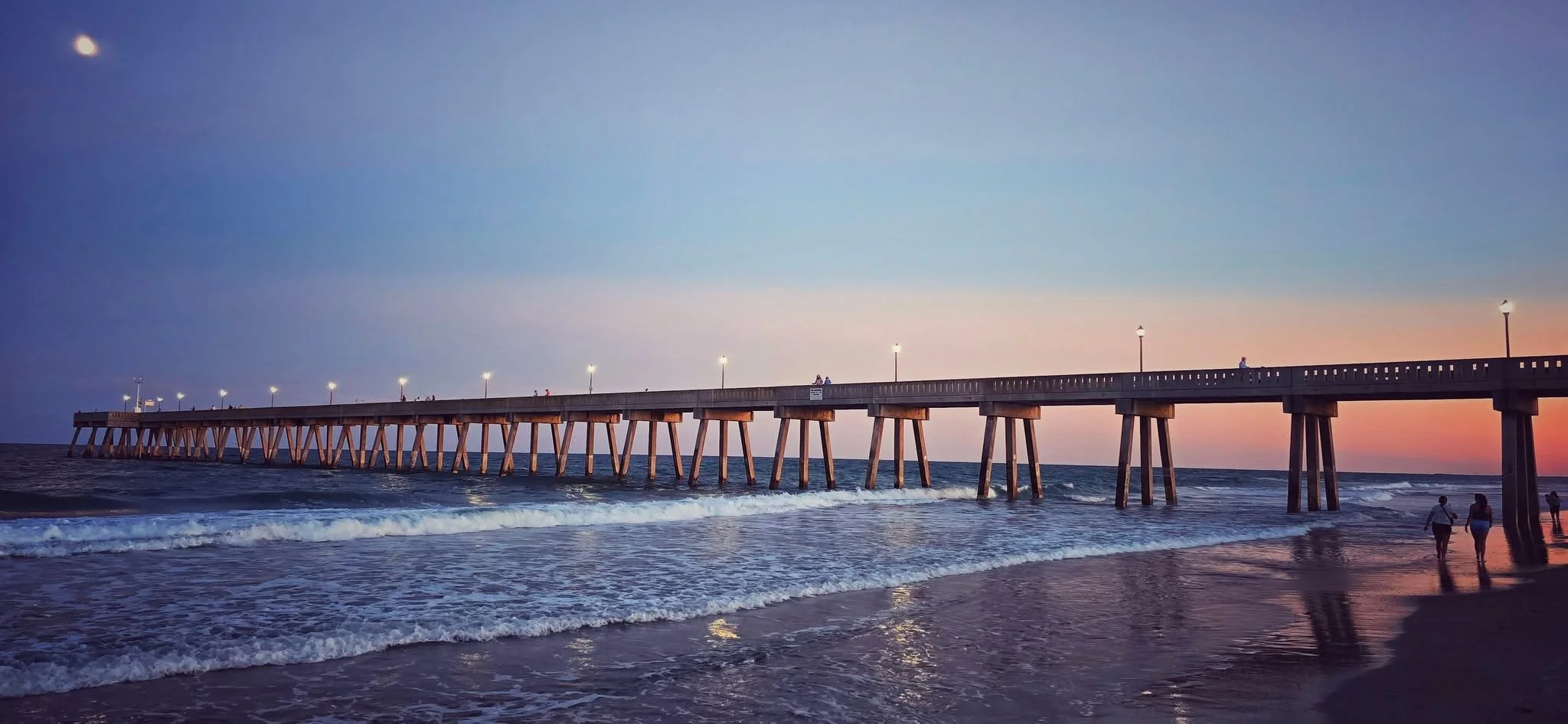 A pier extending into the ocean at sunset, with a few people walking on the pier and along the beach, the sky transitioning from blue to orange.