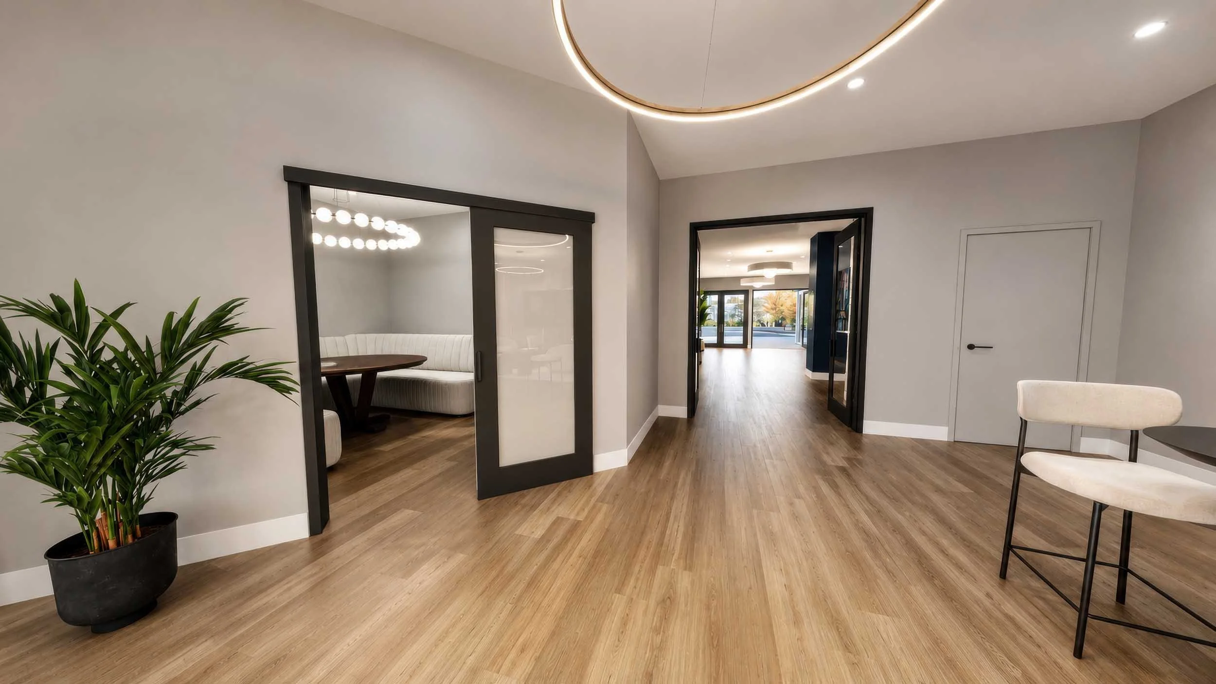 Modern interior space with wooden flooring, white walls, a large potted plant, and seating area visible through sliding glass doors, with additional rooms or areas beyond.