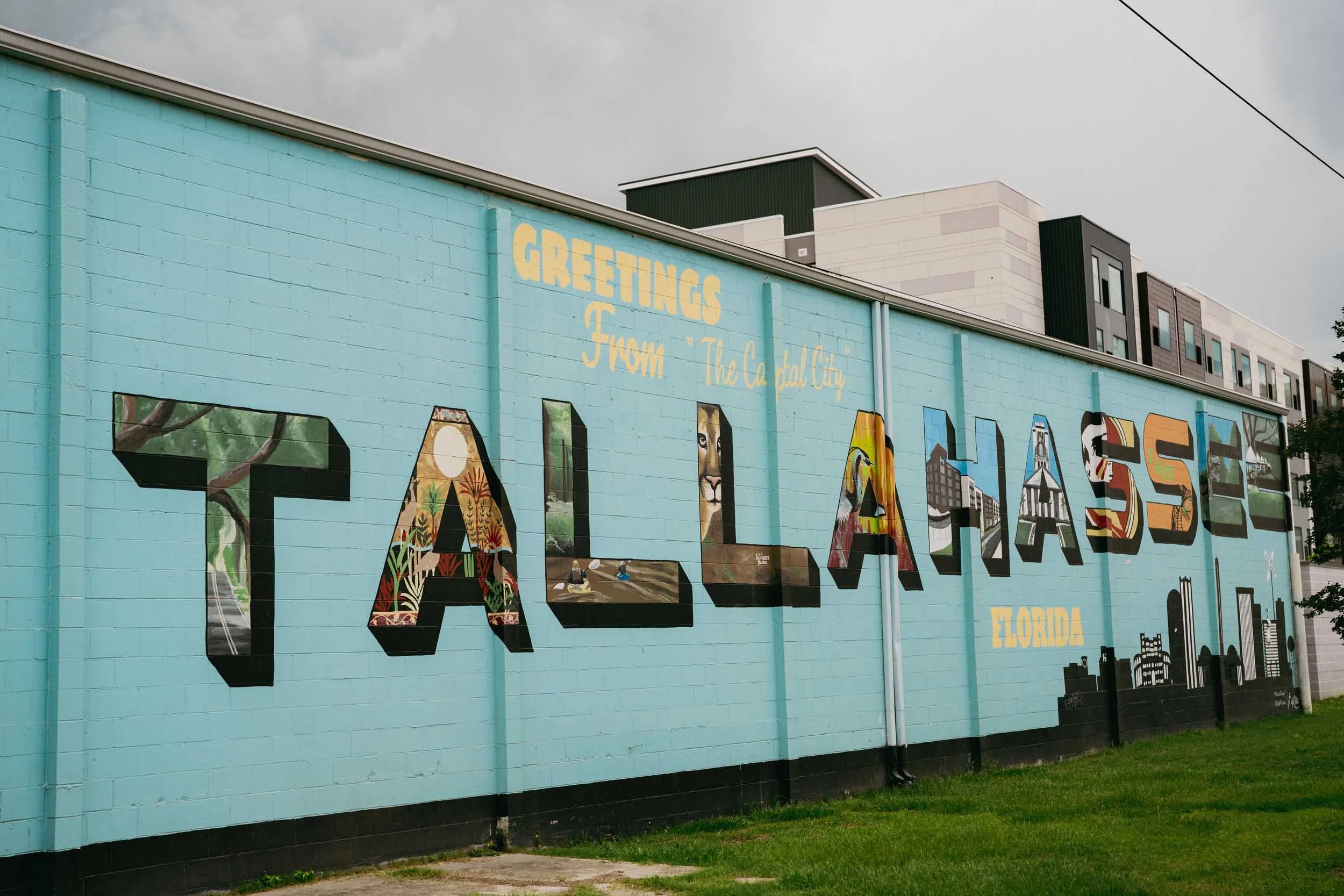 Colorful mural on a blue building wall that spells out "TALLAHASSEE" with each letter featuring different regional landmarks and icons of Florida, with an overcast sky above.