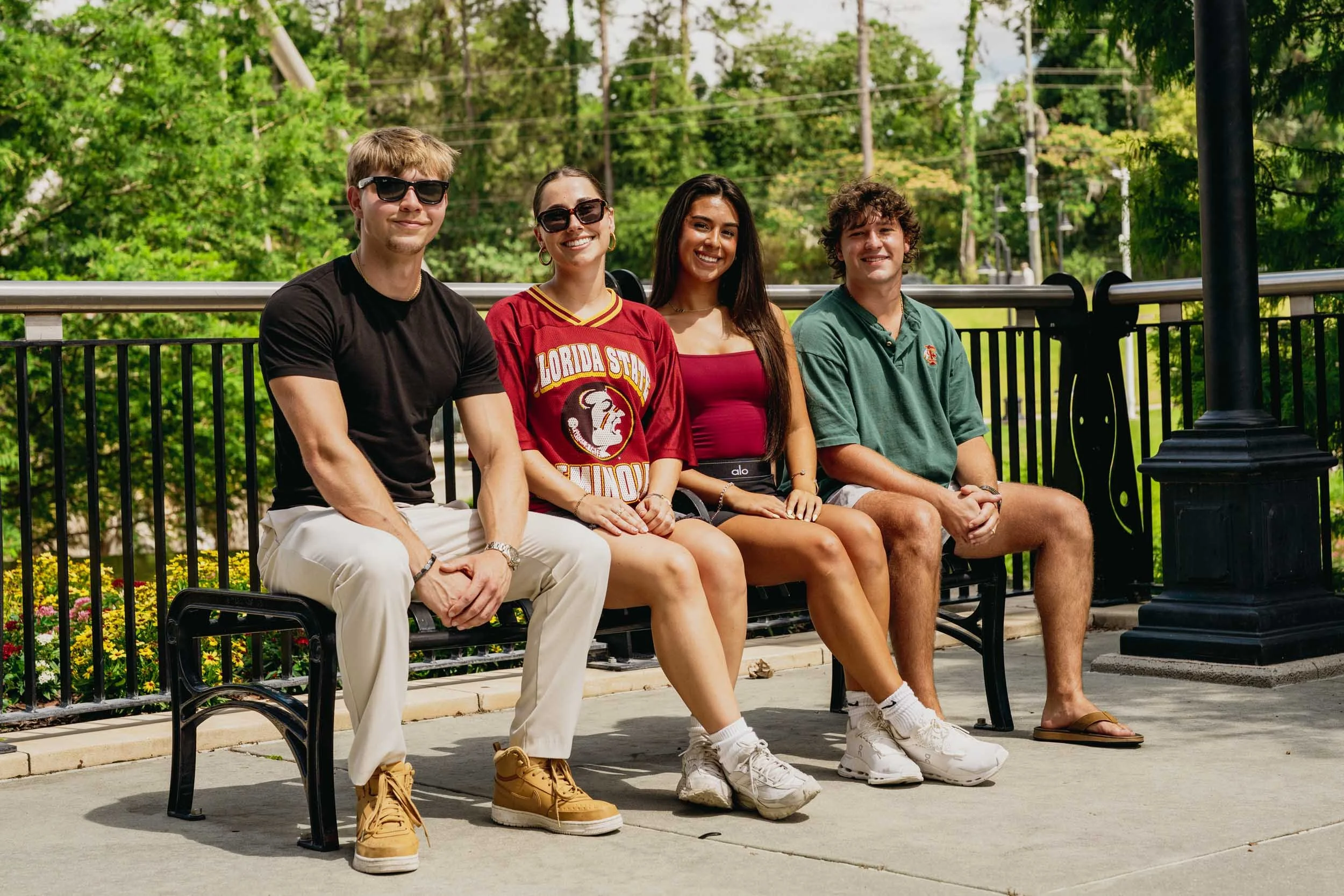 Four young adults sitting on a park bench, smiling, surrounded by greenery and trees, with clothing indicating warm weather.