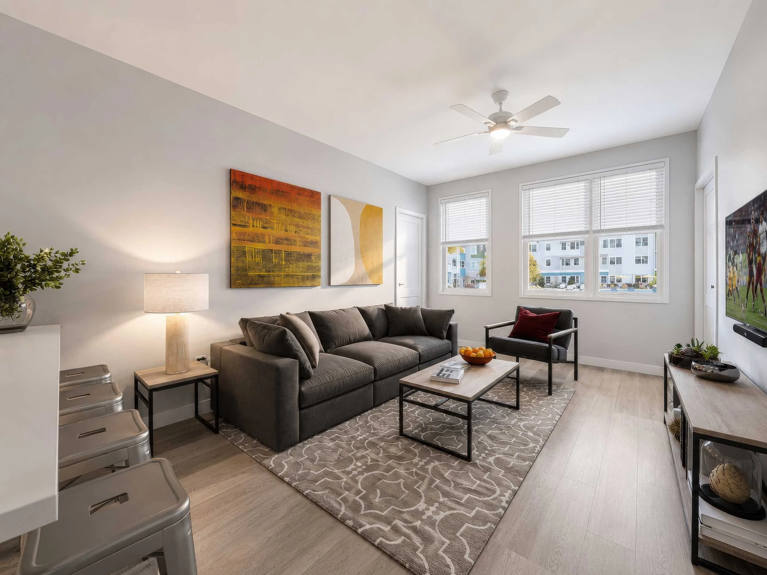 A modern living room with a gray sofa, wall art, a white side table with a lamp, large windows with blinds, a TV, and a ceiling fan.