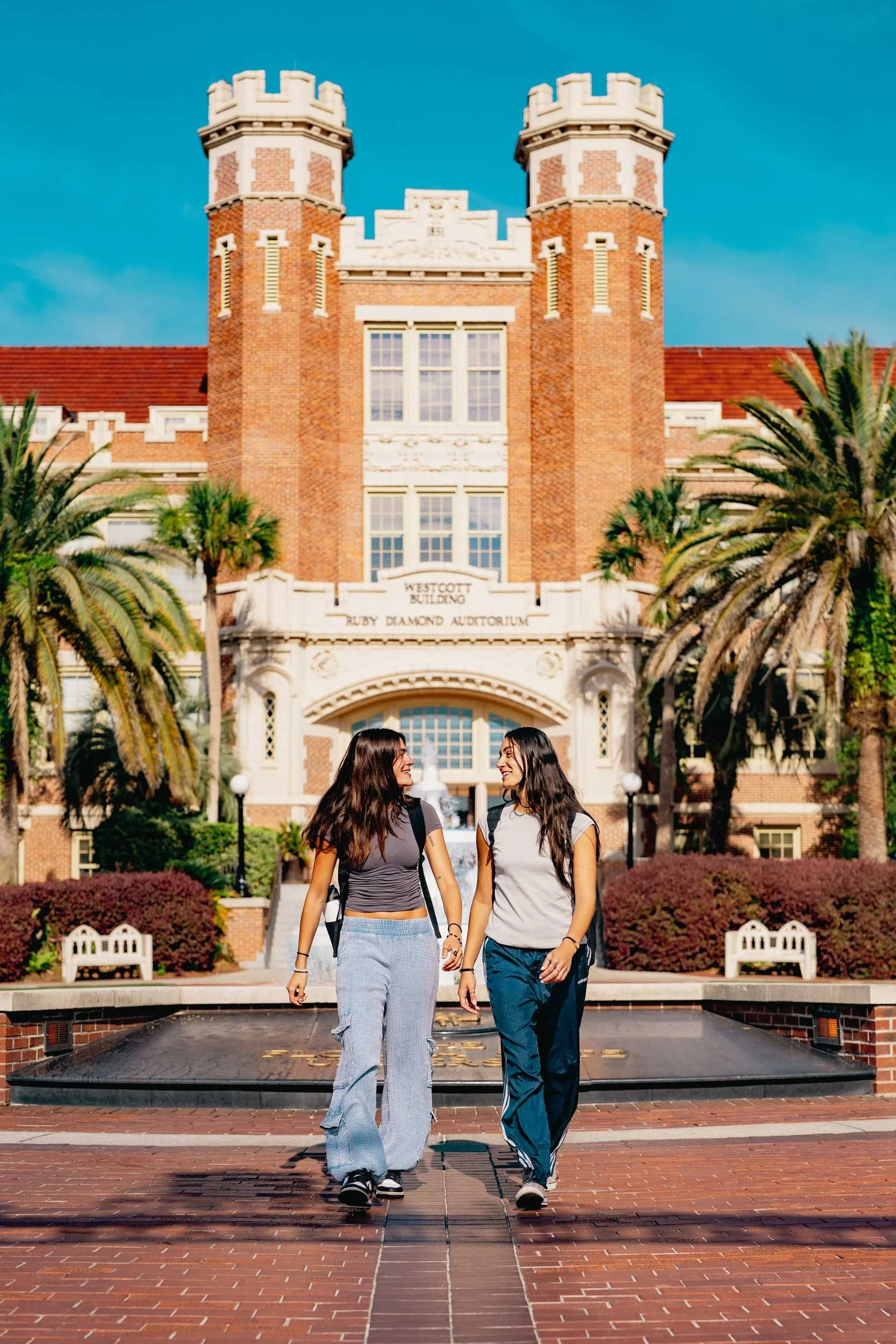 Two young women with backpacks walking together in front of a historic brick building with towers, palm trees, and a fountain.