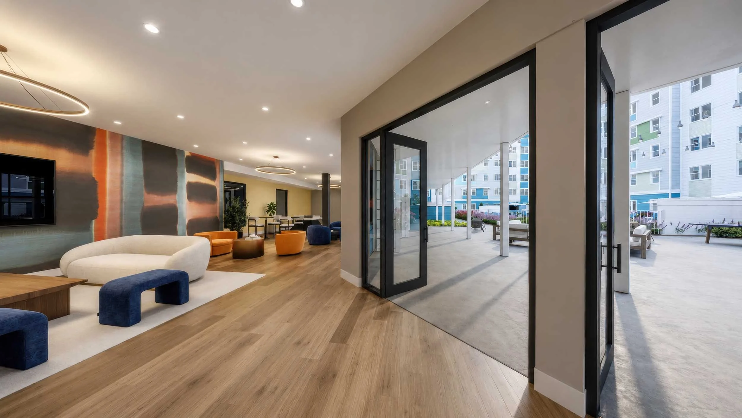 Modern indoor living space with wooden floors, colorful furniture, and large glass doors leading to an outdoor patio in a high-rise building.