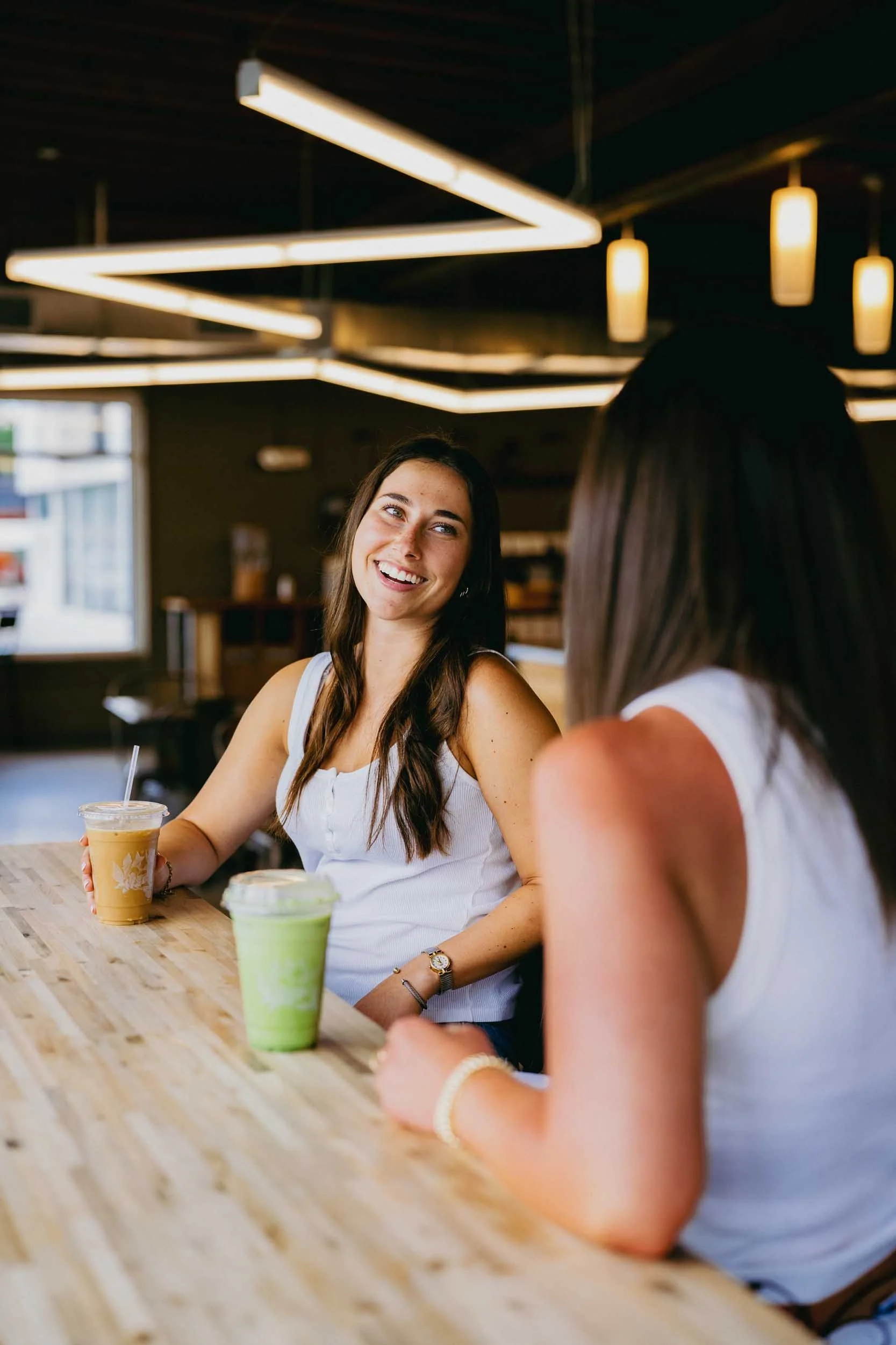 Two young women sitting at a wooden table in a cafe, smiling and talking. The woman facing the camera has long dark hair and is holding a drink in a clear plastic cup. The other woman, with her back to the camera, has long dark hair. There are two smoothies on the table, and the cafe has modern lighting fixtures.