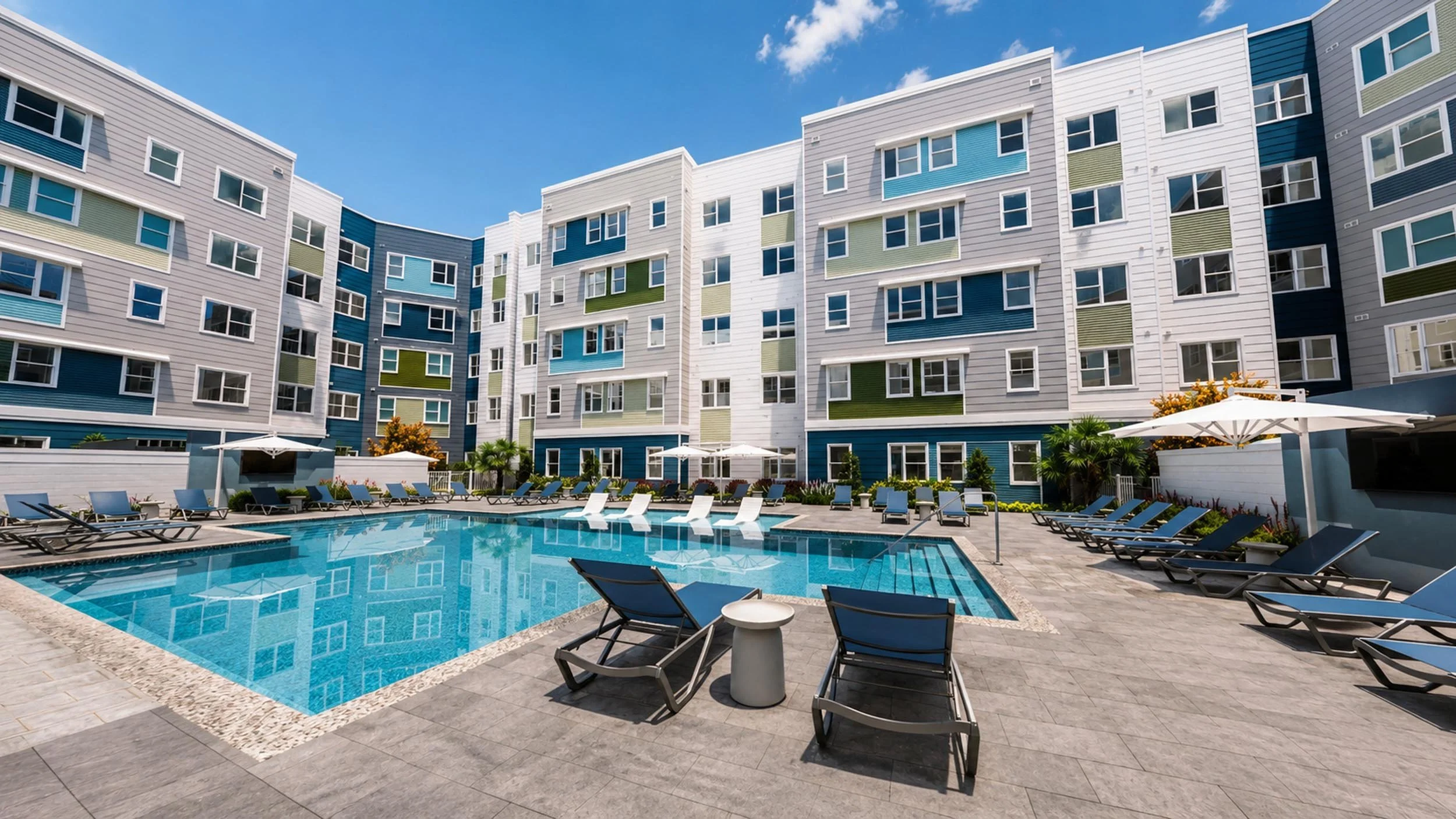 Modern apartment complex with colorful exterior, swimming pool surrounded by lounge chairs, umbrellas, and lush greenery under a blue sky.