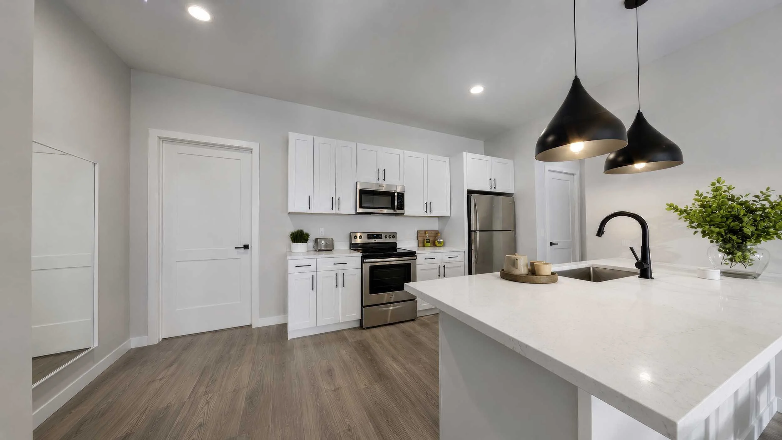 Modern white kitchen with stainless steel appliances, black pendant lights, a green plant, and a white countertop with cups.