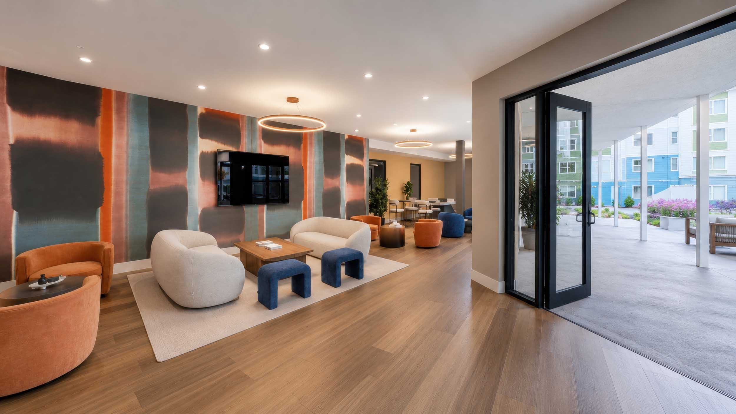 Modern lounge with a contemporary design, featuring colorful armchairs, a mounted flat-screen TV, and decorative circular ceiling lights. Large sliding glass doors open to an outdoor patio with a view of nearby apartment buildings.