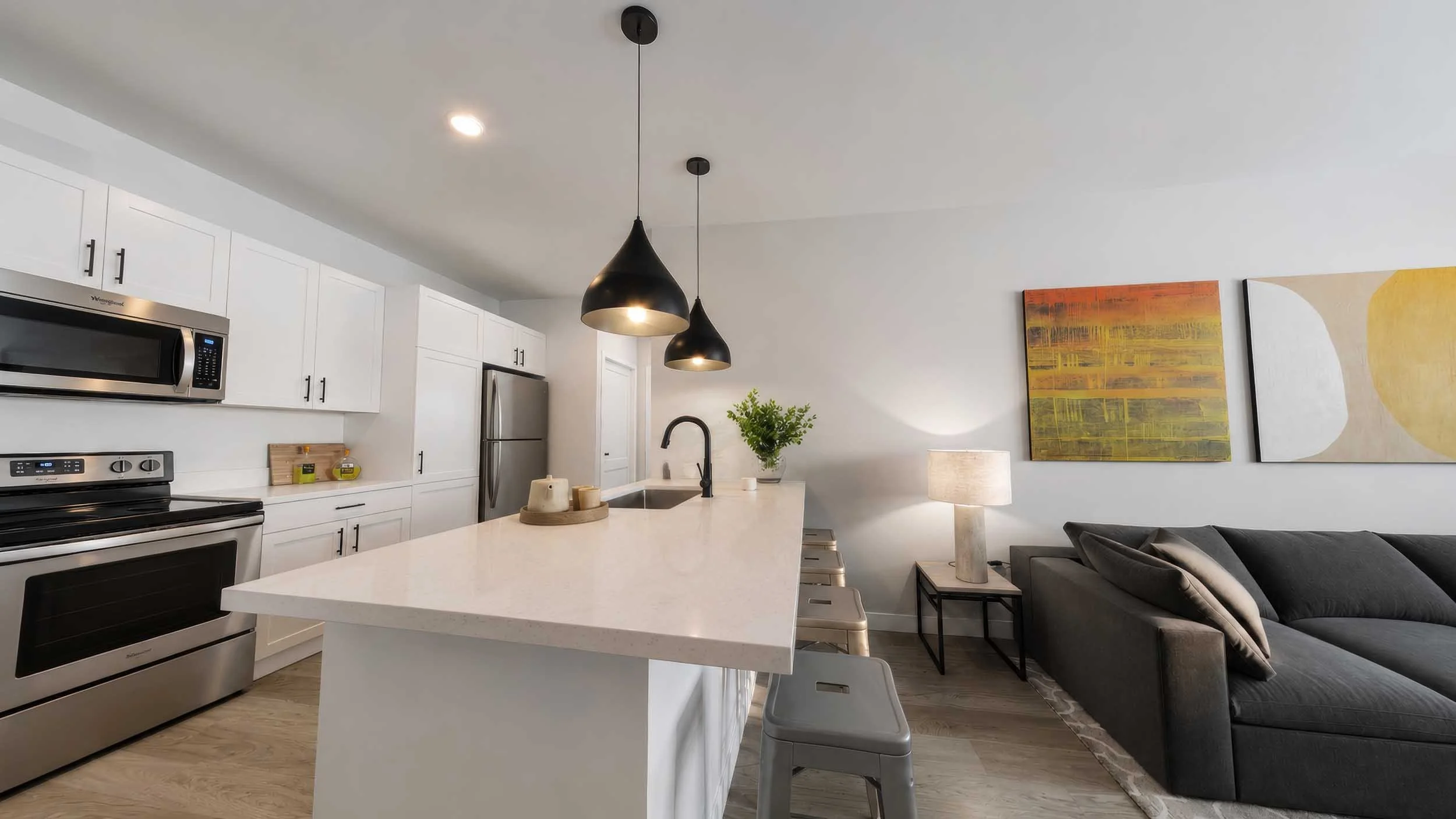 Modern kitchen with white cabinets, stainless steel appliances, black pendant lights above a white island, and artwork on the wall with a gray sofa nearby.