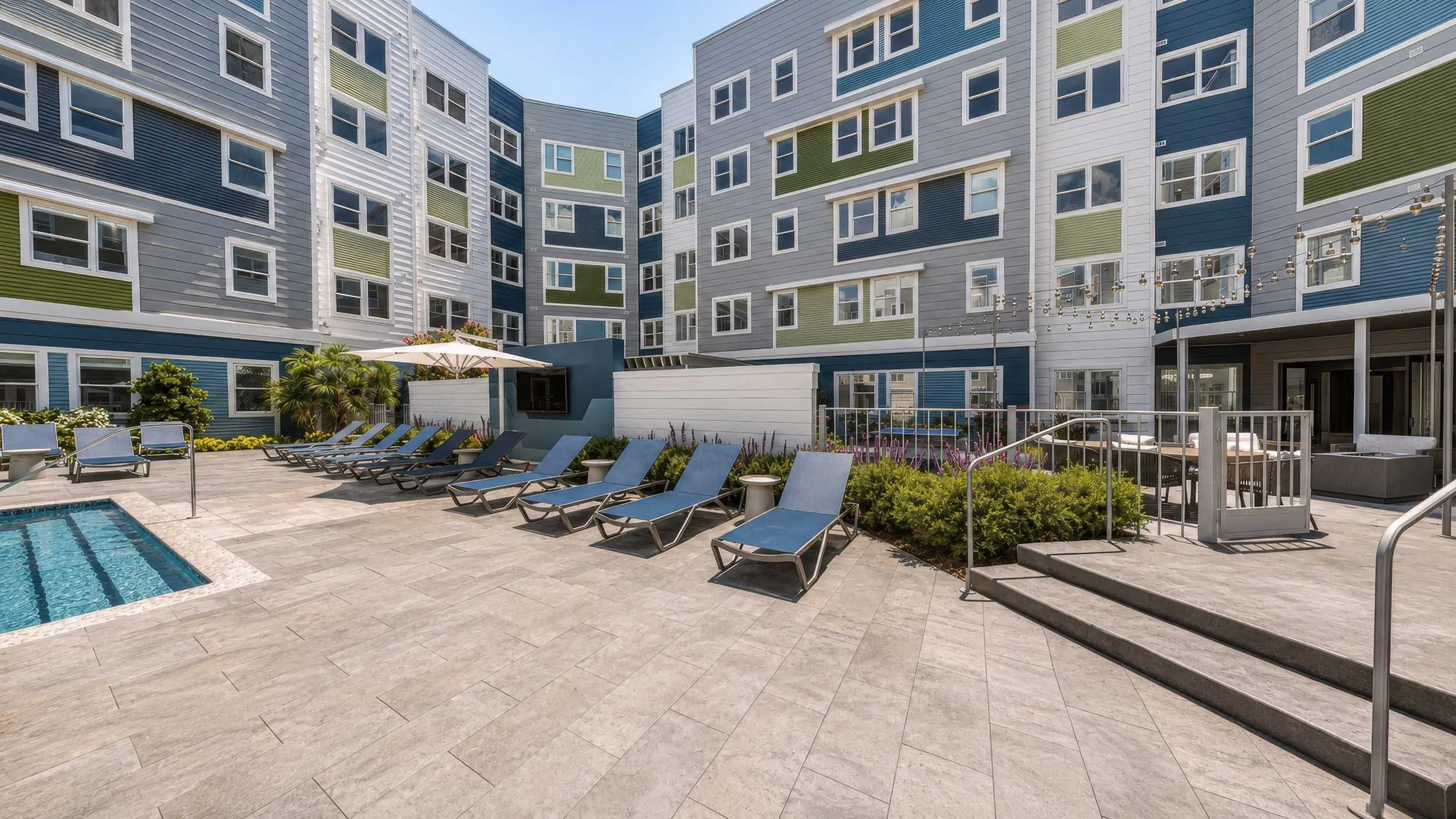 View of a modern apartment complex courtyard with a swimming pool, lounge chairs, greenery, and a patio area.