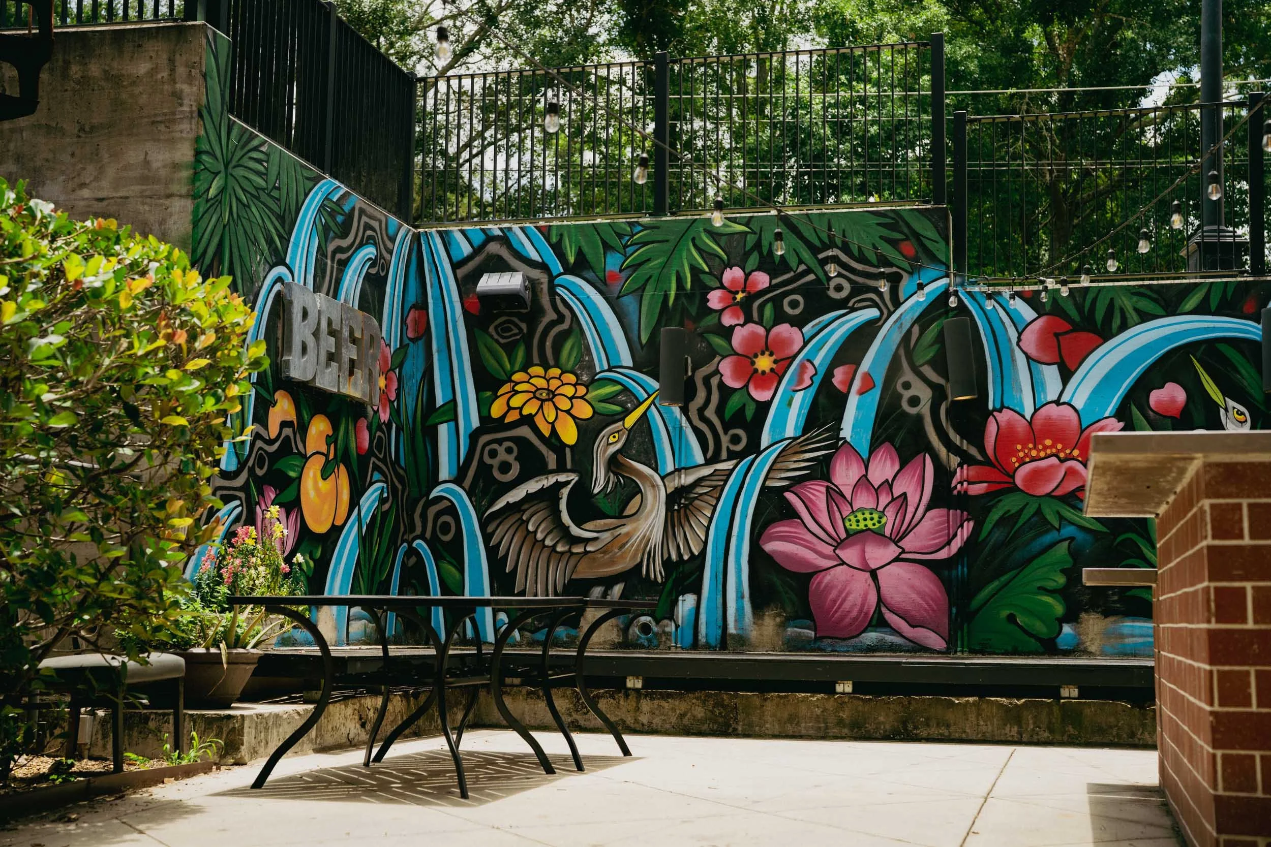 Colorful mural with flowers, water, and fierce animal faces, situated on a brick wall in an outdoor patio area with trees. String lights hang above.