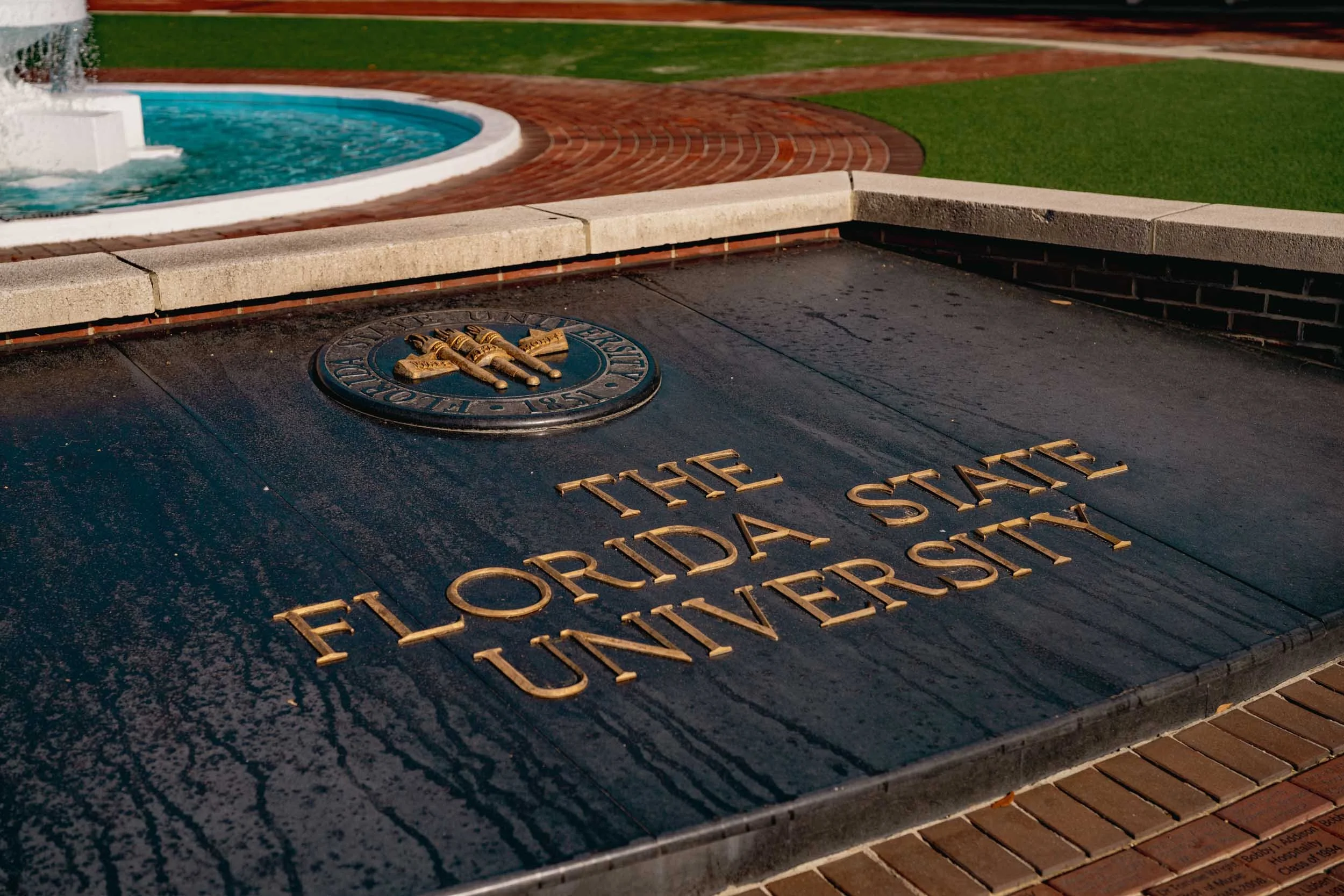 Close-up of a memorial plaque with a statue of flags on it, located outdoors near a water fountain with a brick and grass background.