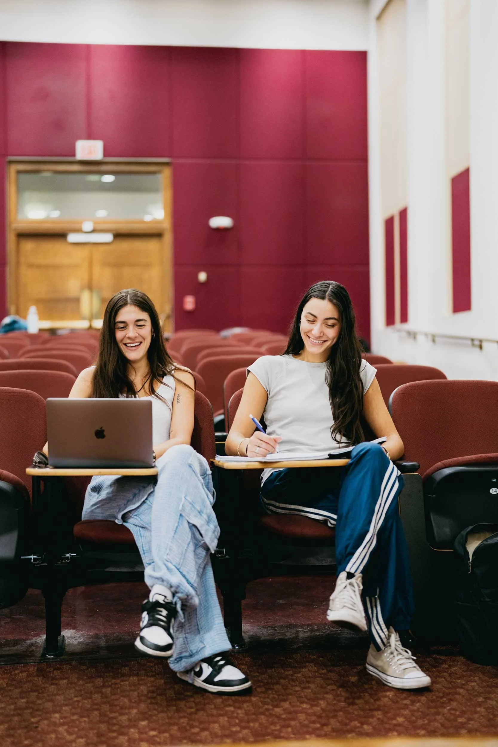 Two young women sitting in an auditorium, smiling, with a laptop and notes in front of them.