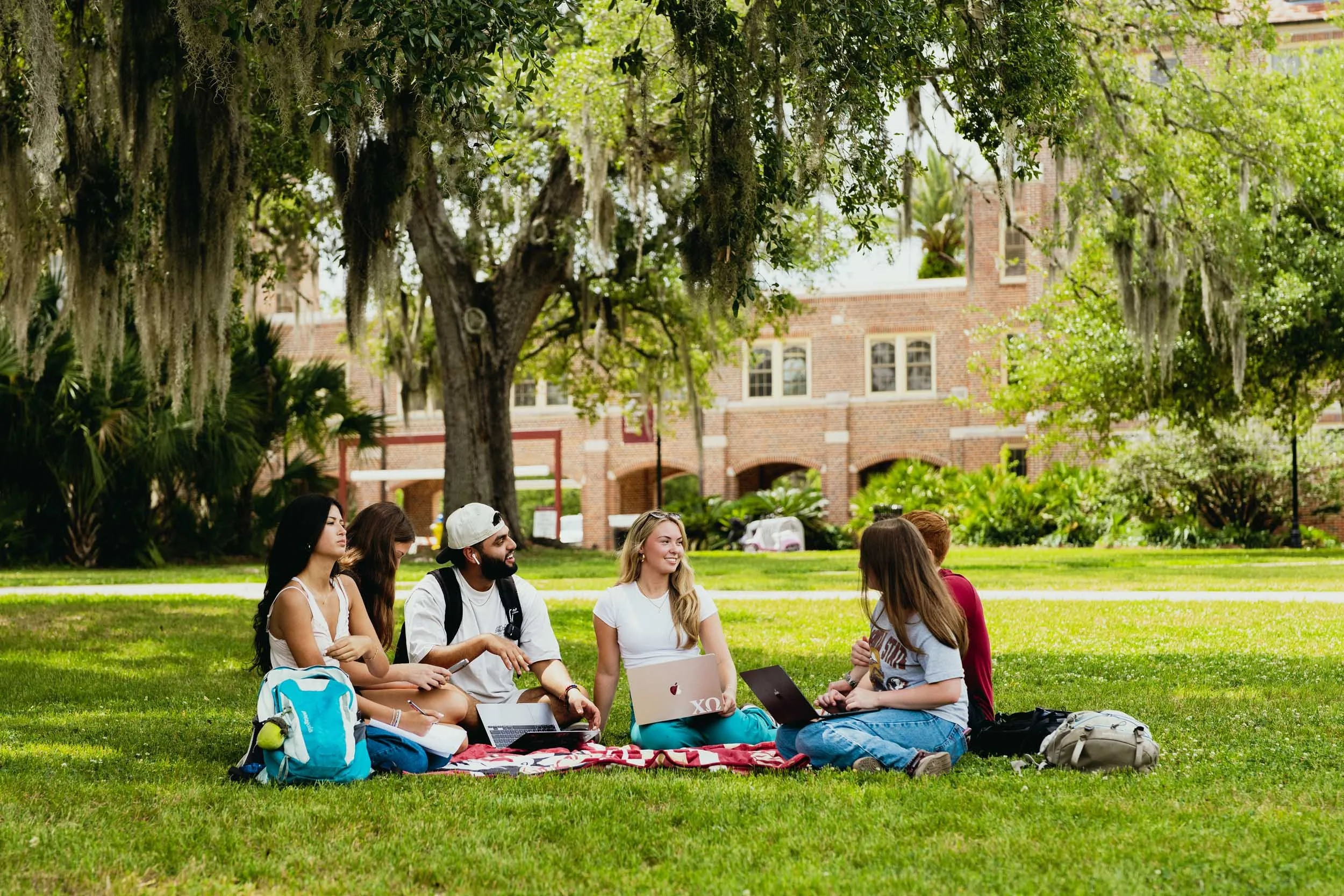 A group of young people sitting on the grass in a park, surrounded by trees, with a brick building in the background, some laptops and backpacks around them.