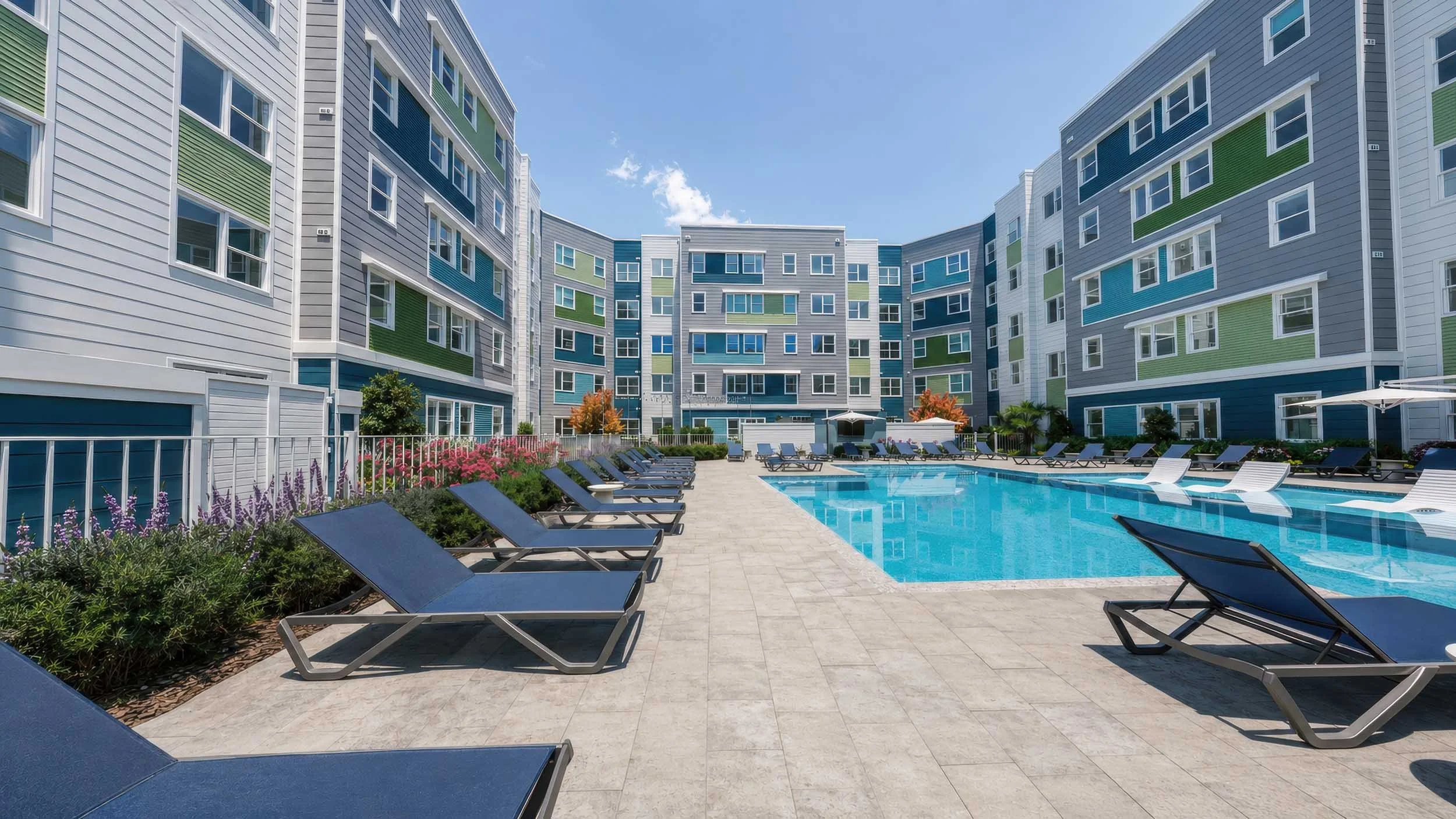 A modern residential apartment complex with a swimming pool area surrounded by lounge chairs, colorful landscaping, and multi-story buildings with blue, green, gray, and white exterior.